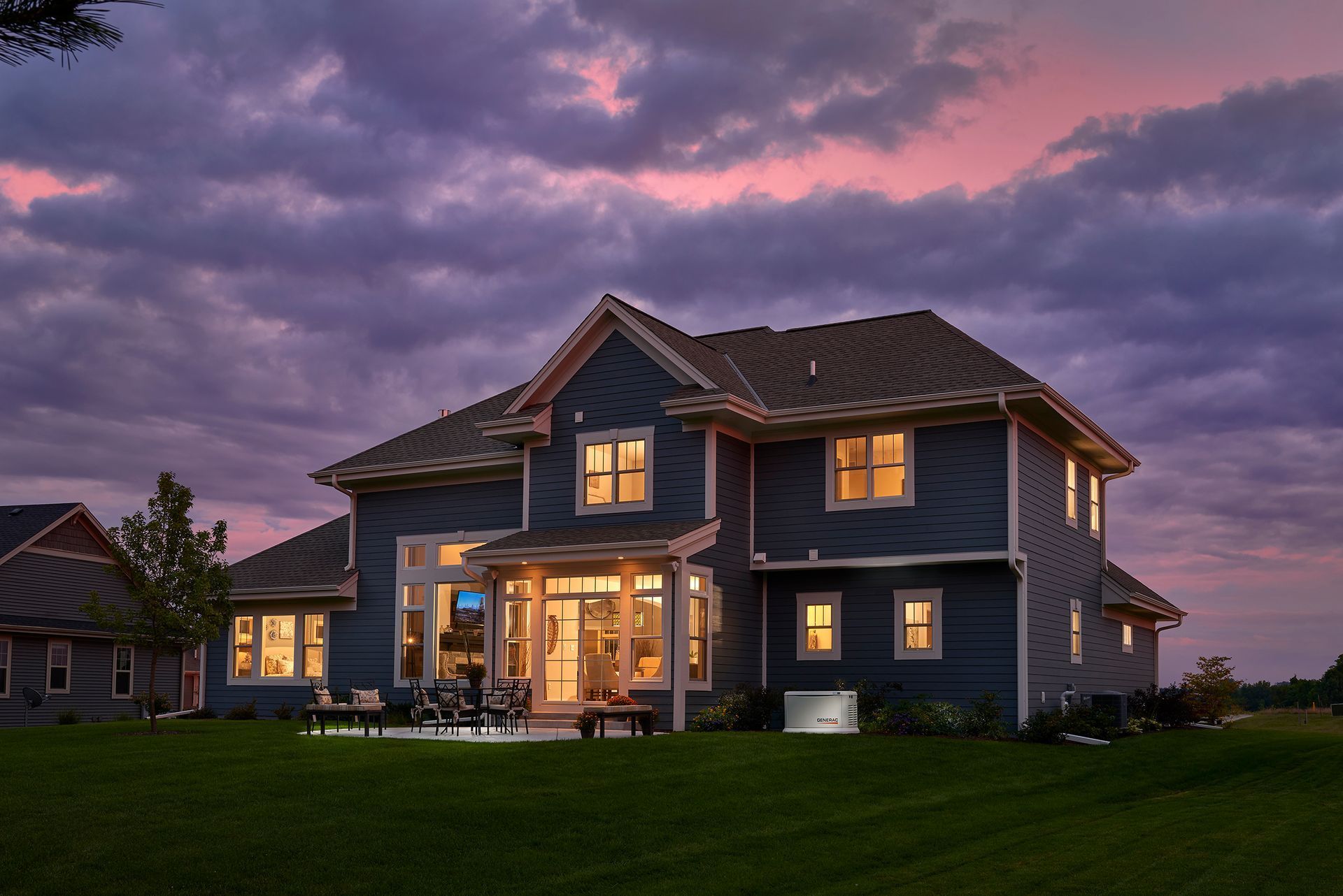Two-story gray house with warm lights glowing at dusk under a purple cloudy sky
