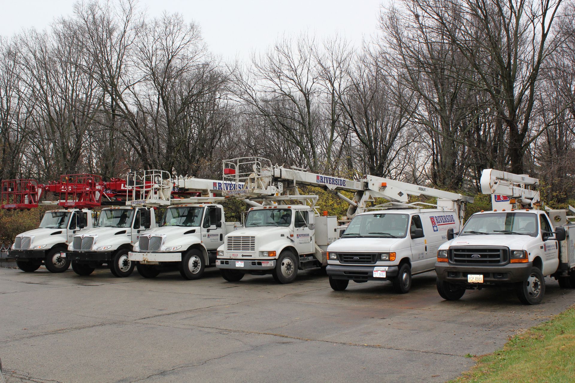 Line of utility trucks and bucket lifts parked in a lot with leafless trees behind them