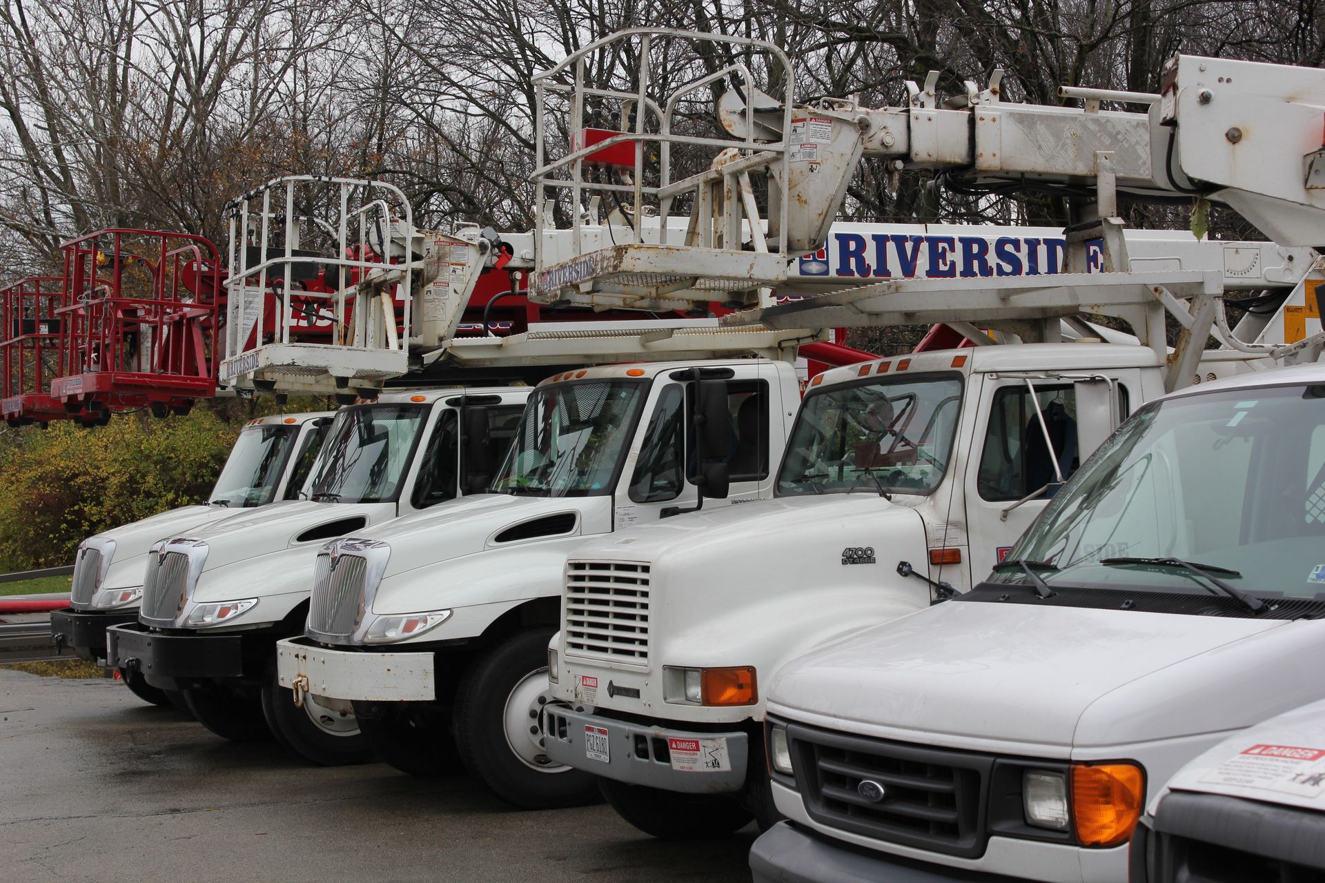 White utility trucks and a red boom truck parked in a lot on a cloudy day, with bare trees behind them