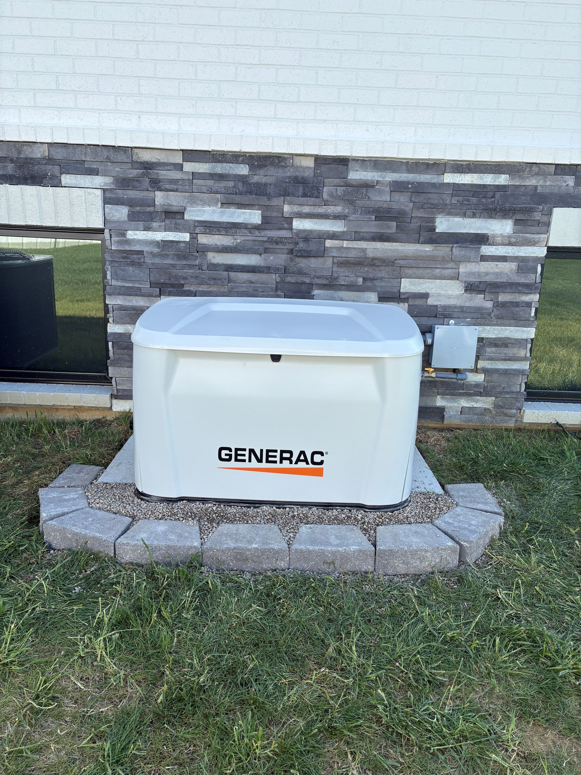 White Generac backup generator on a stone pad against a house wall and grass lawn