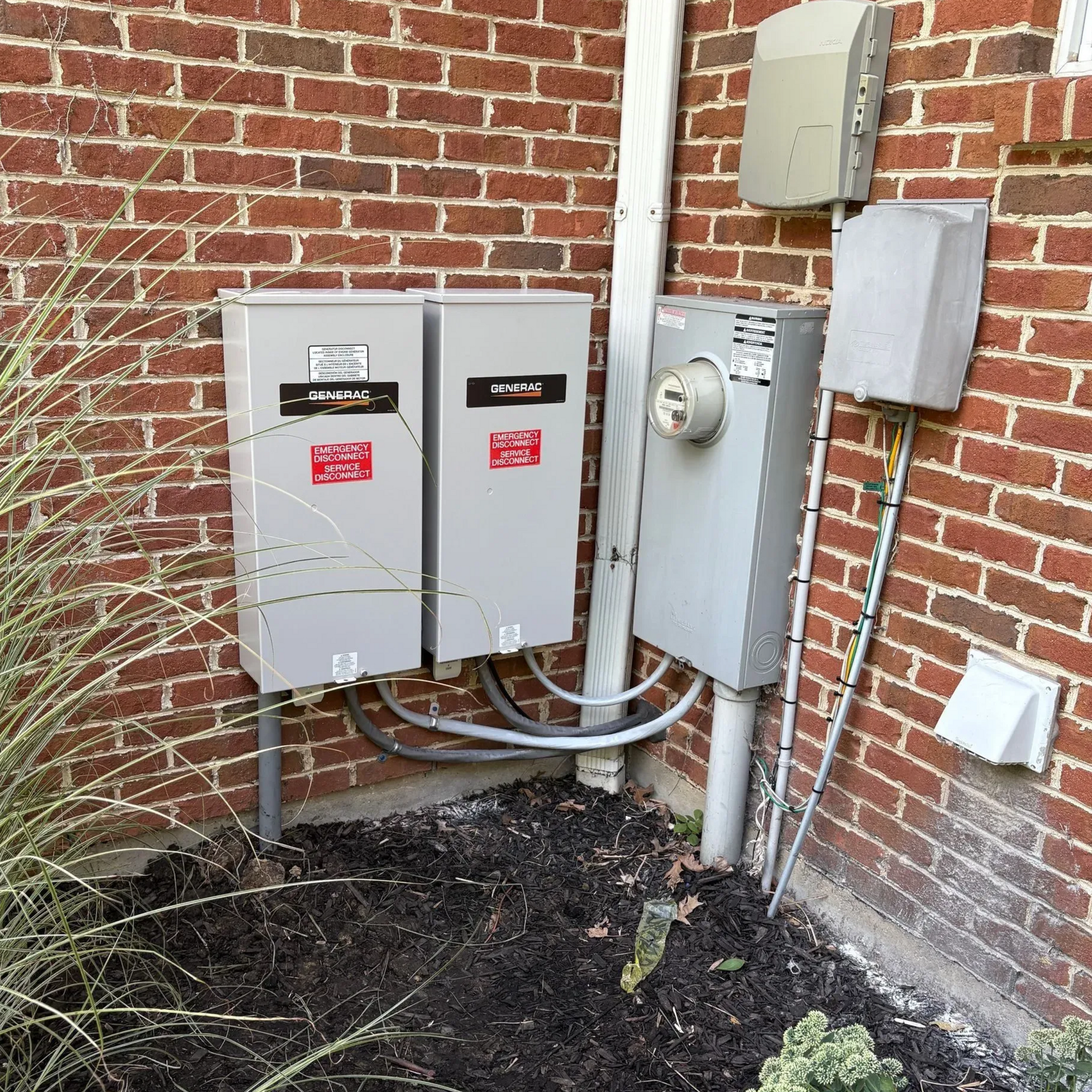 Electrical utility boxes and conduit mounted on a brick wall beside a building foundation
