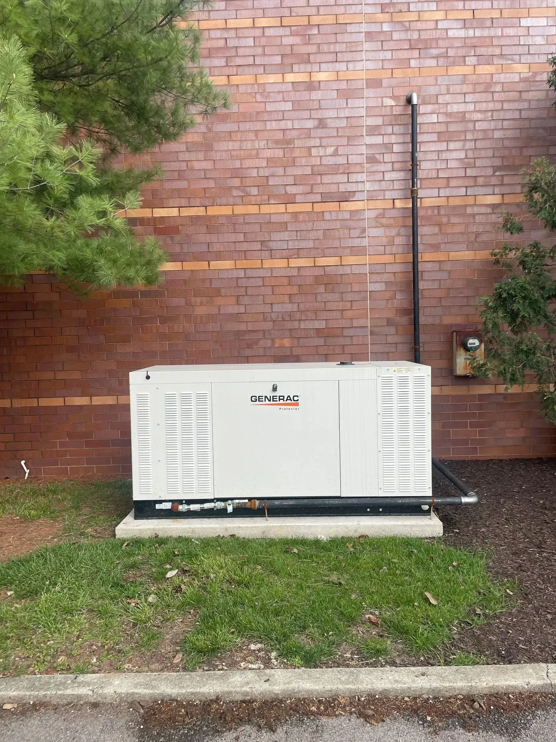 White HVAC unit beside a brick wall, with grass and a tree in the foreground