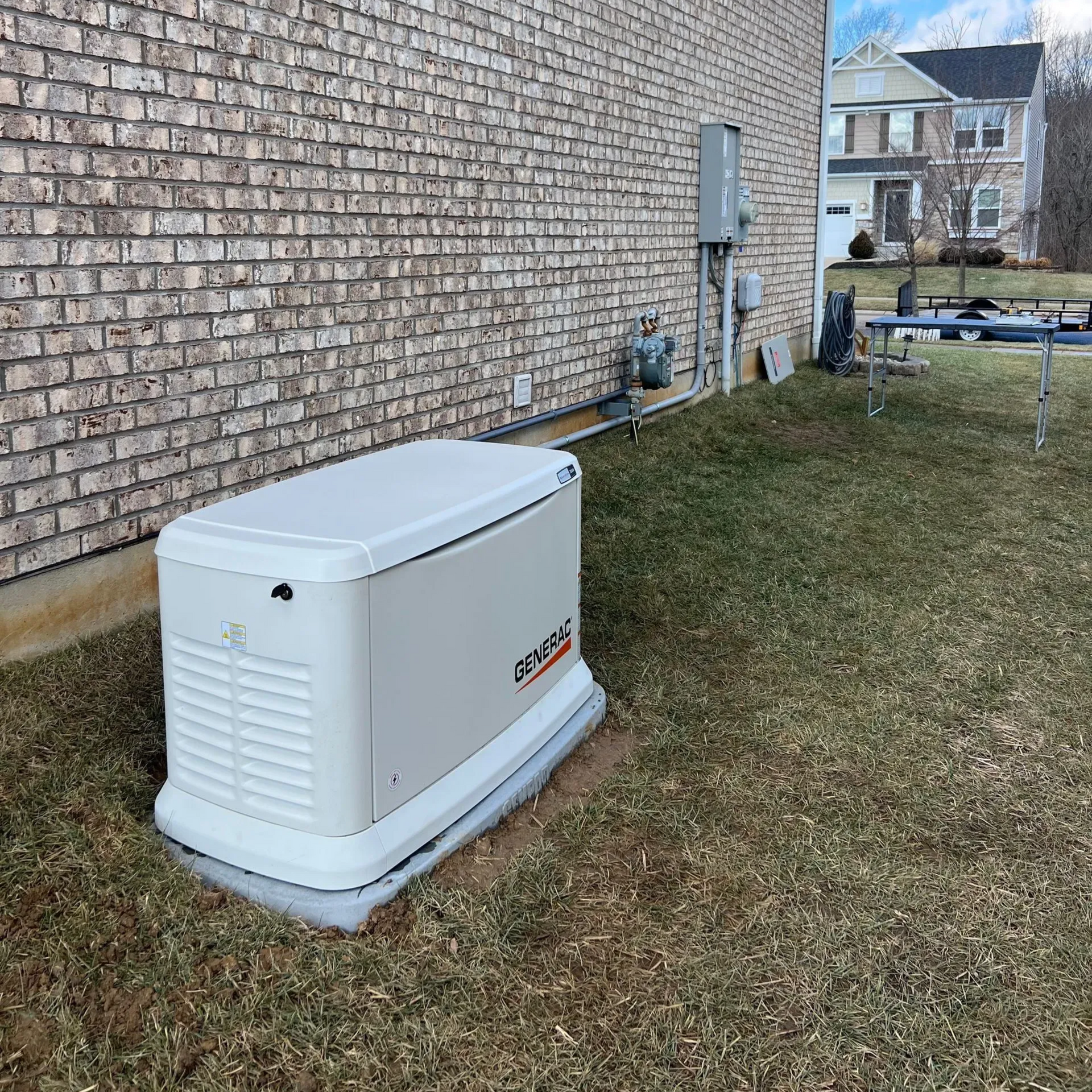 White standby generator beside a brick house on a grassy yard, with neighboring homes in the background