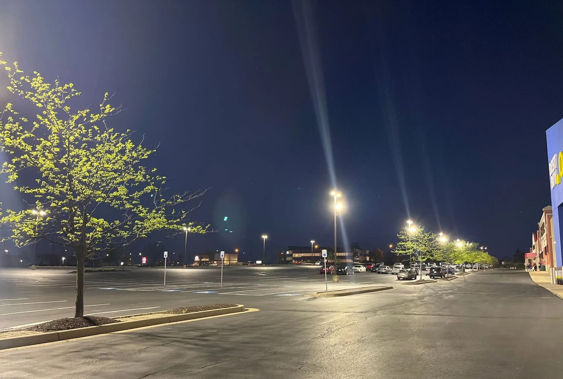 Empty parking lot at night with bright lights, a lit tree, and buildings along the right side.