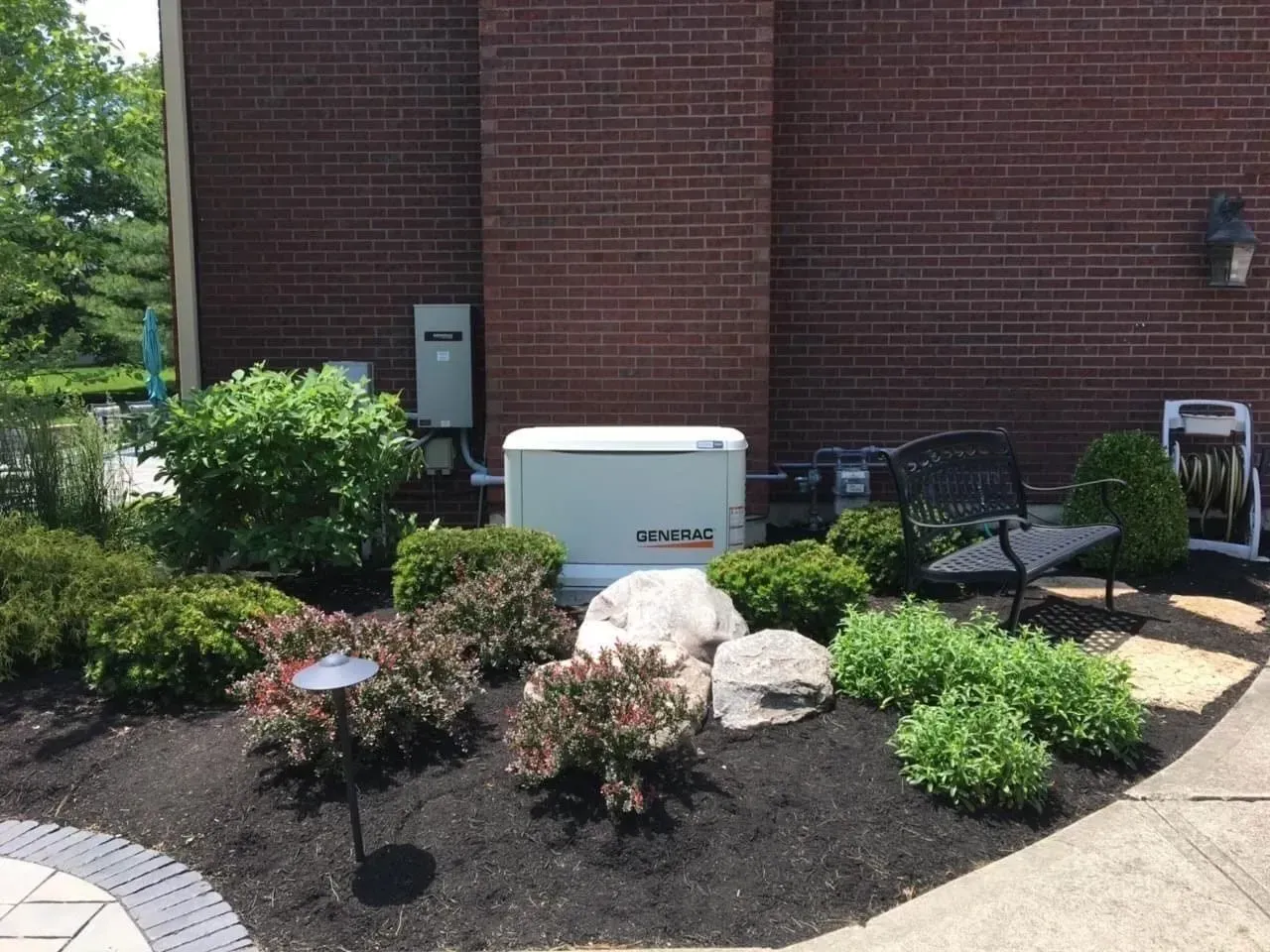 Front of a brick building with shrubs, a white utility box, and outdoor chairs on a concrete patio.