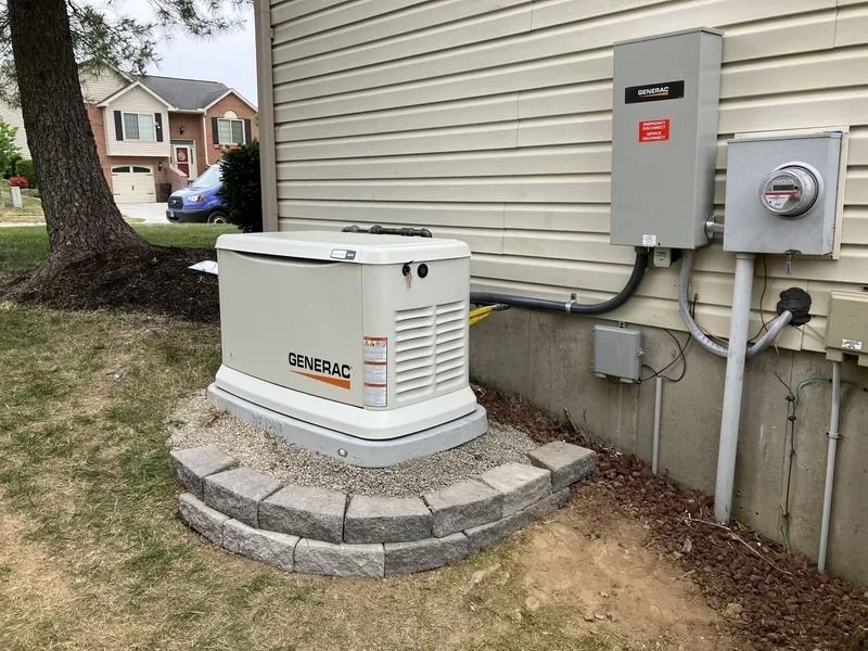 Outdoor standby generator beside a house, mounted on a stone pad next to electrical panels.