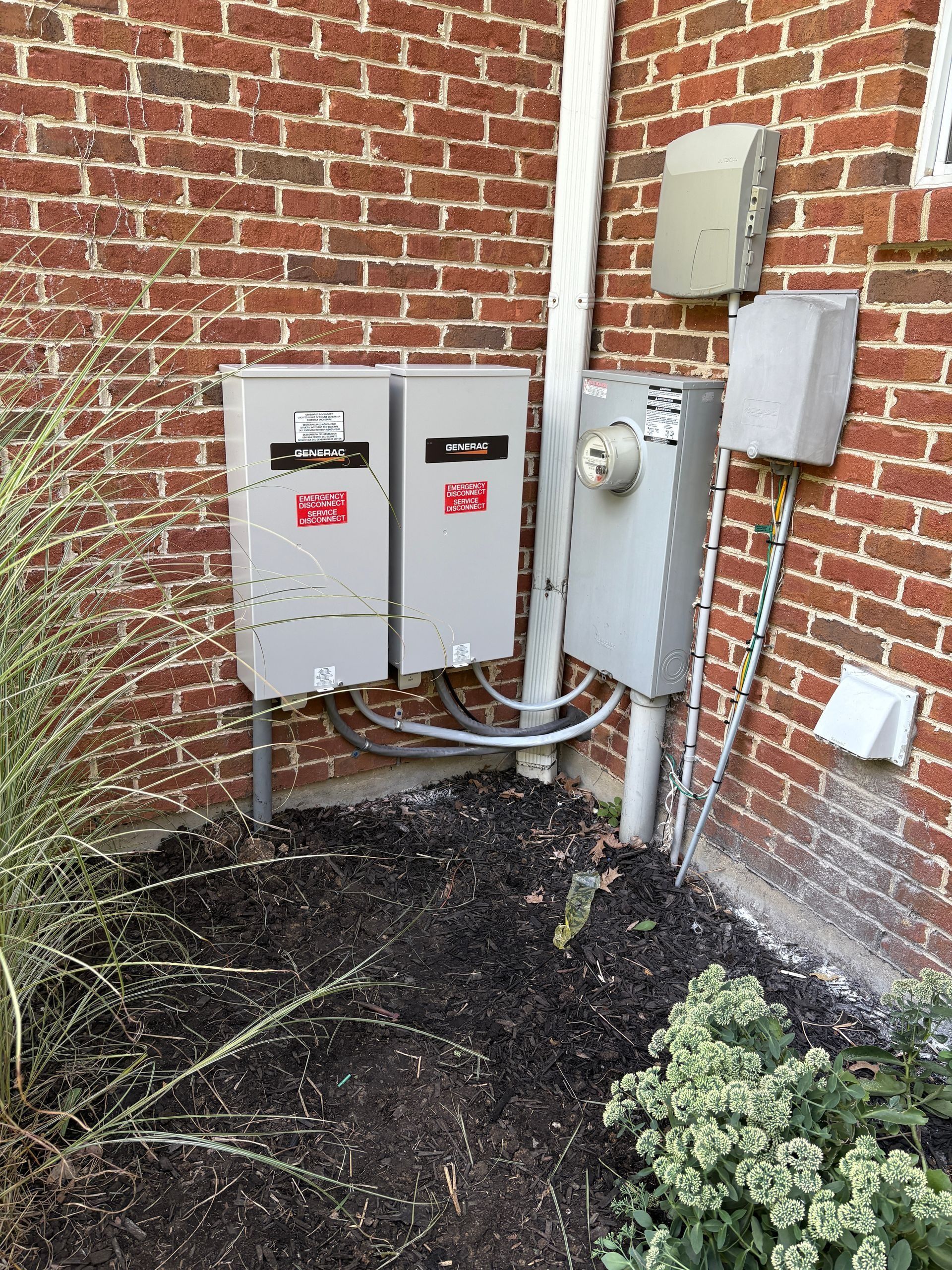 Outdoor electrical meters and utility boxes mounted on a brick wall beside conduit and weeds.