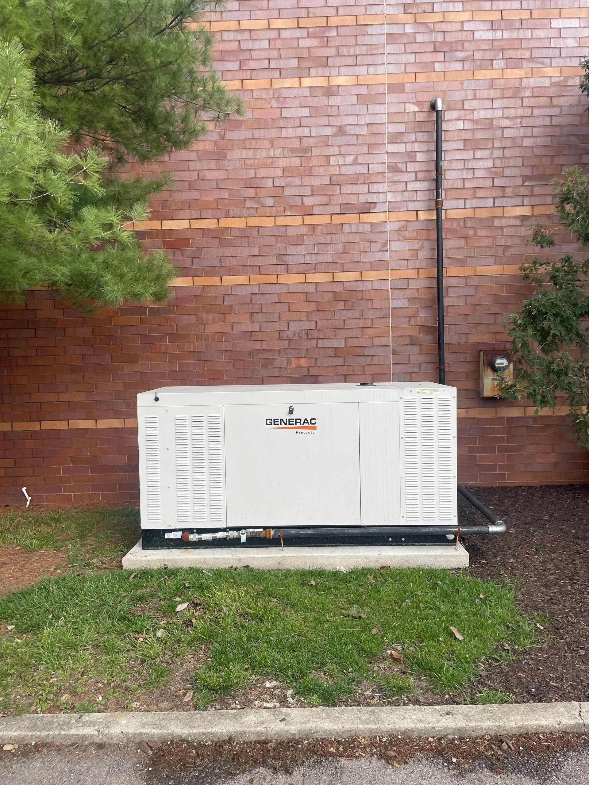 White generator unit on grass beside a brick wall, with a tree and utility pipes nearby.