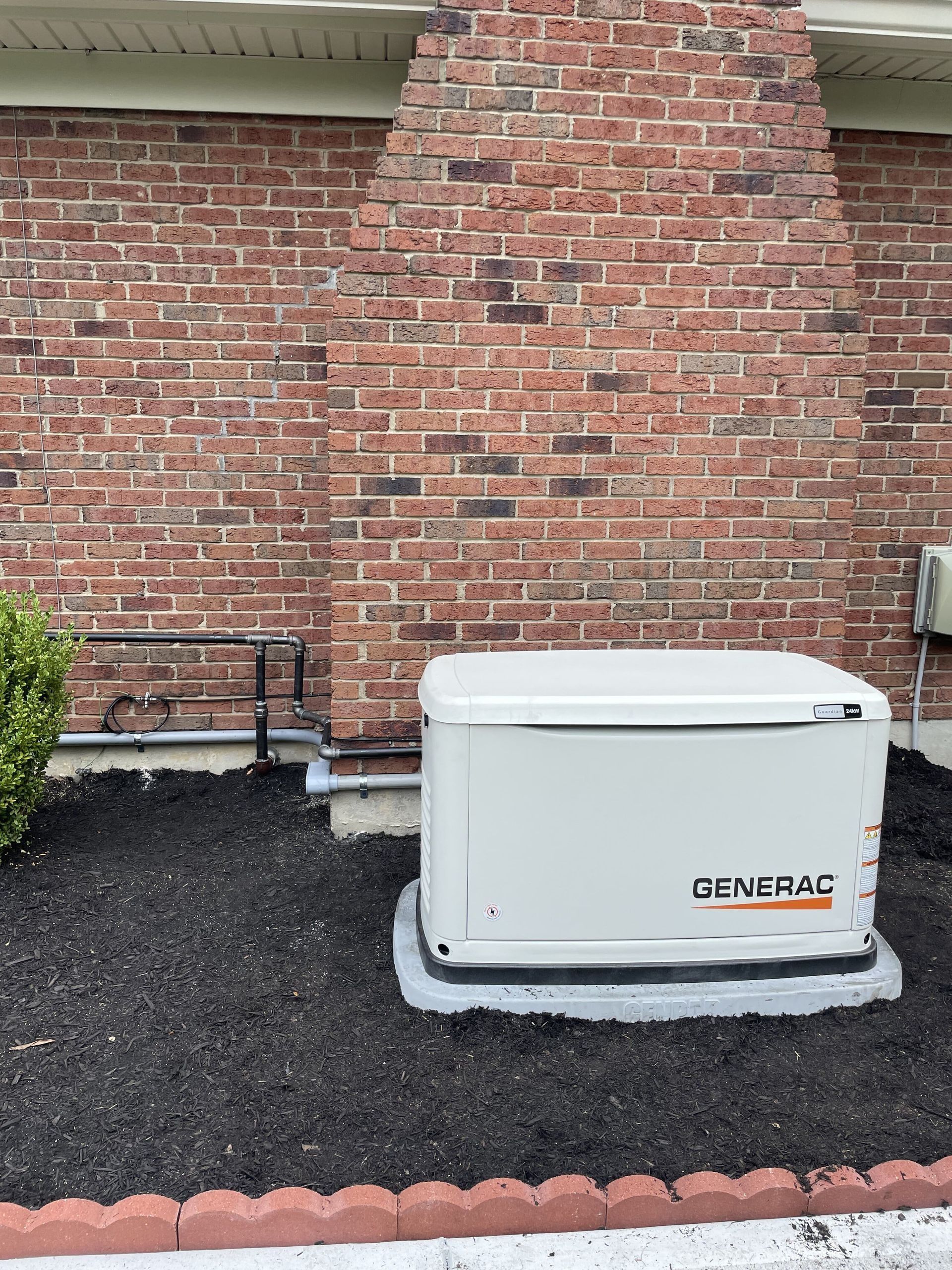 White generator on gravel beside a brick house wall and small bush.