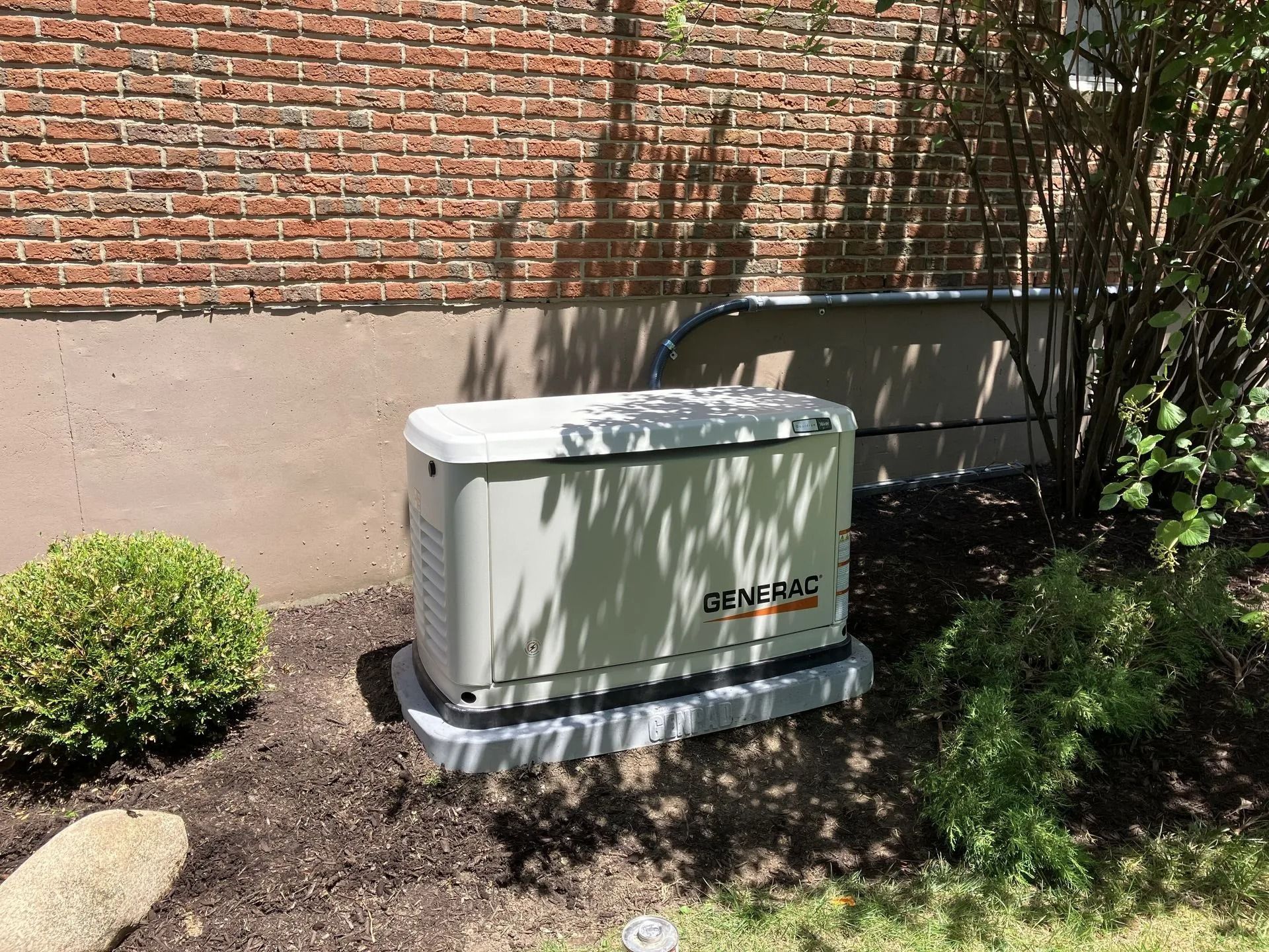 Outdoor HVAC unit beside a brick wall, partly shaded by a tree, with shrubbery nearby.