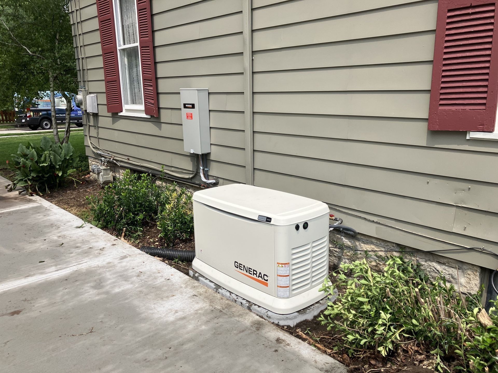 White generator unit beside a house on a concrete pad, near shrubs and a utility meter