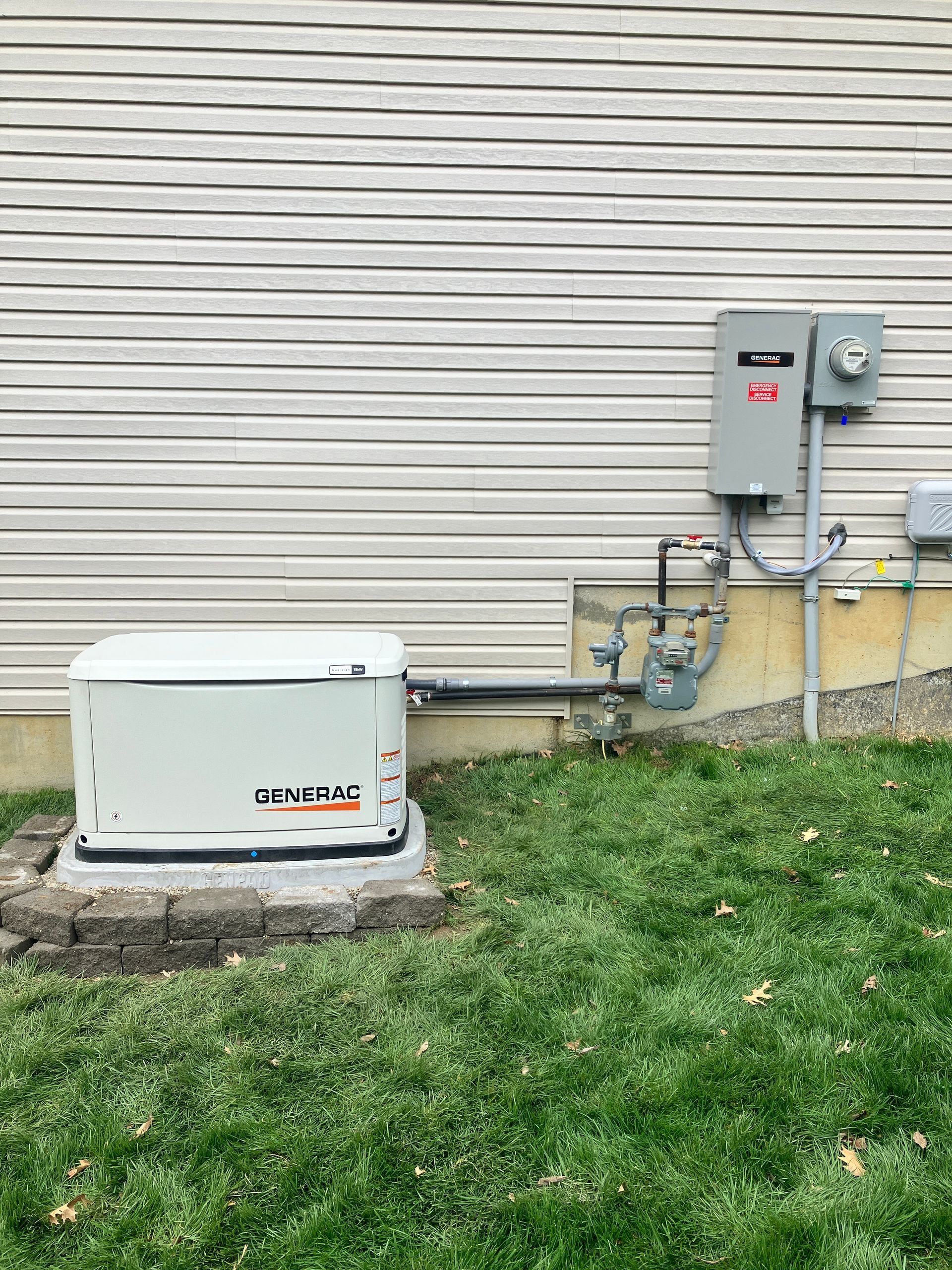 Outdoor utility equipment beside a house: white generator, gas meter, and electrical box on concrete pads.