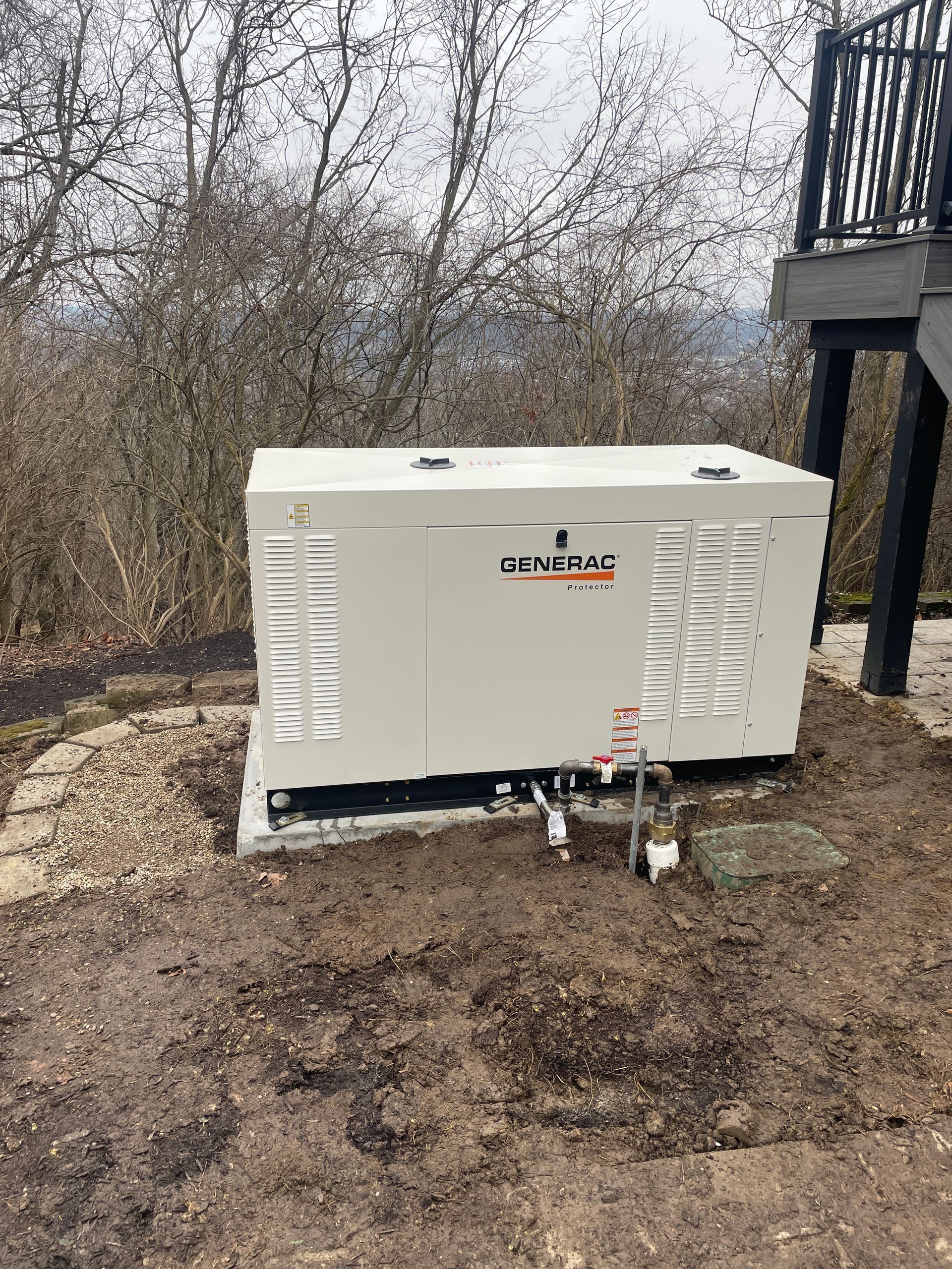Outdoor generator unit beside a house, with pipes and dirt ground in a wooded yard.