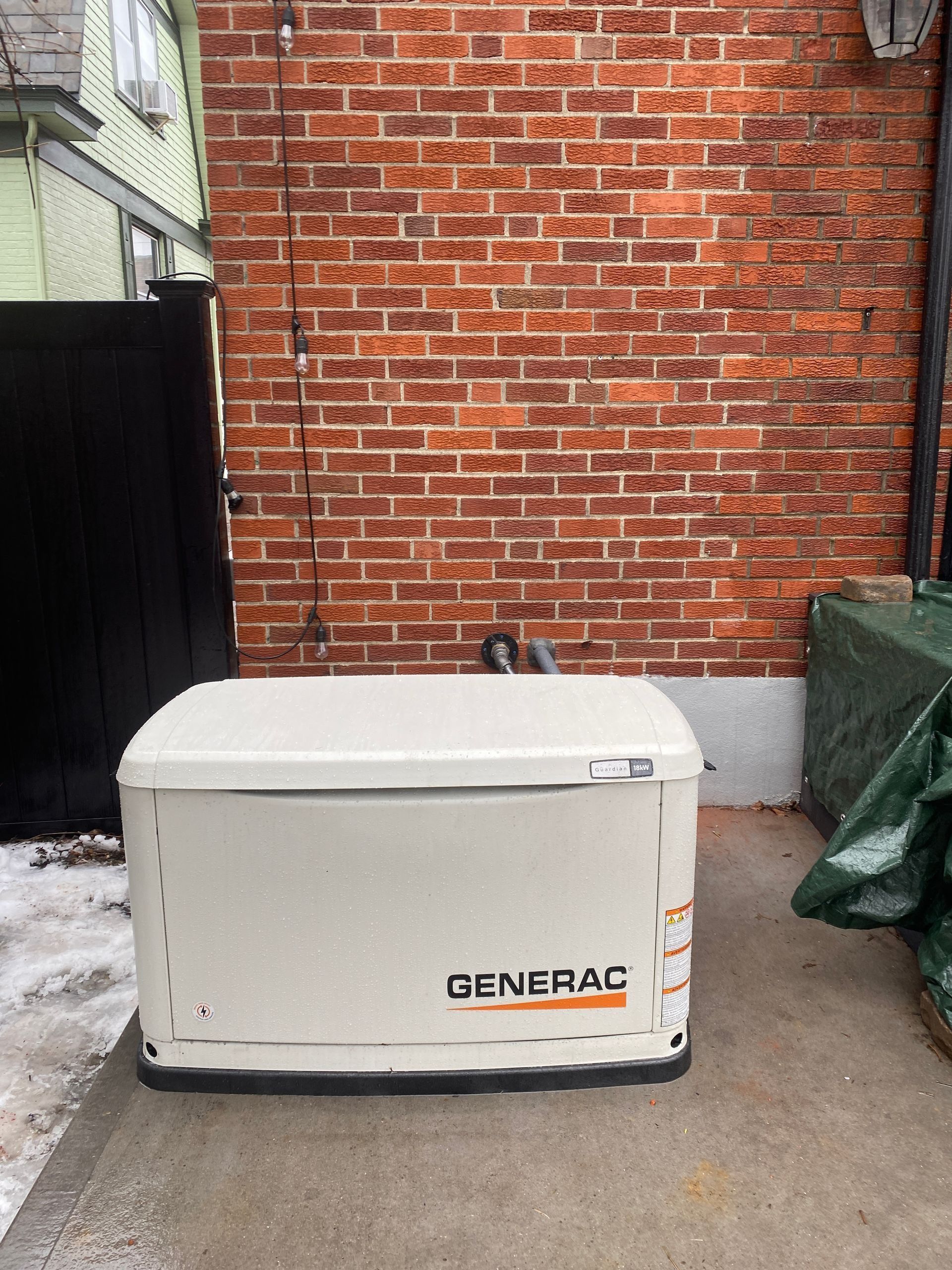 White Generac generator outside a brick building, beside a black fence and green bins.