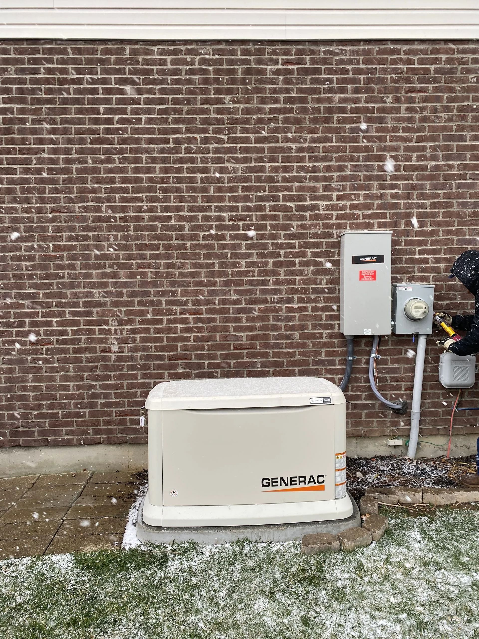 Beige generator unit beside electrical panels against a brick wall outdoors.