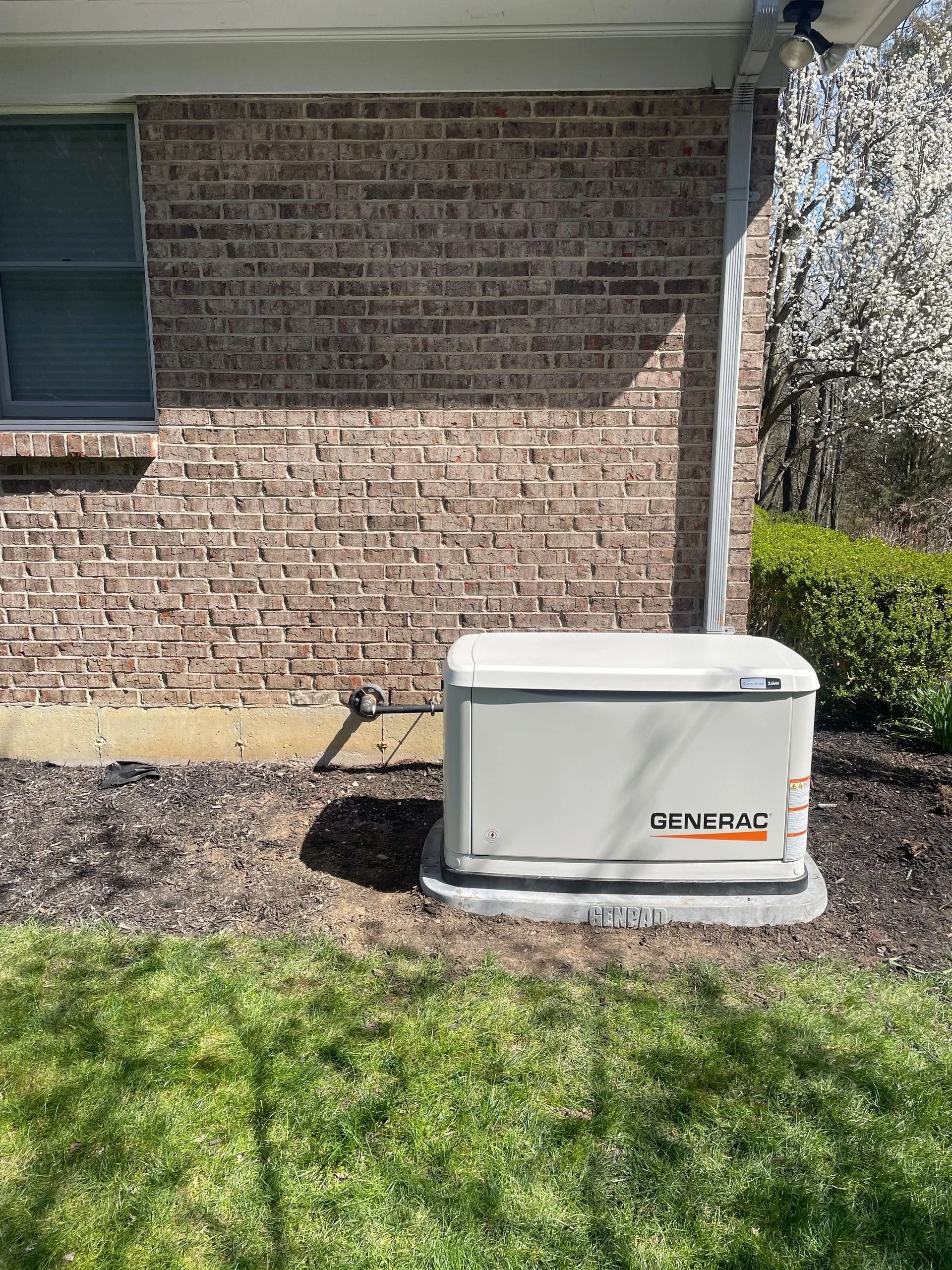 Outdoor generator beside brick house in sunny yard, with white cover and orange logo.