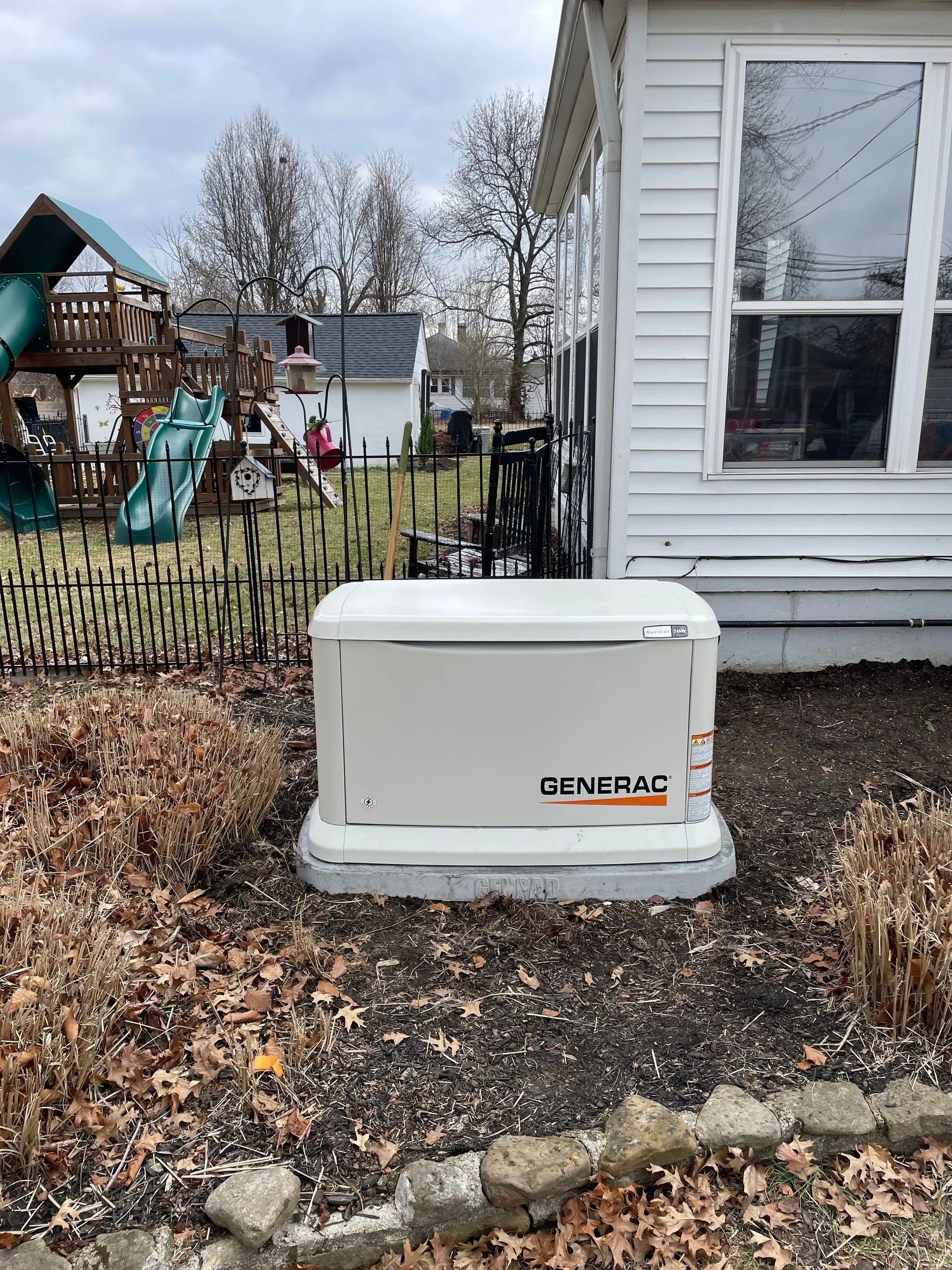 White Generac generator beside a house in a leaf-covered yard, with a playground in the background.