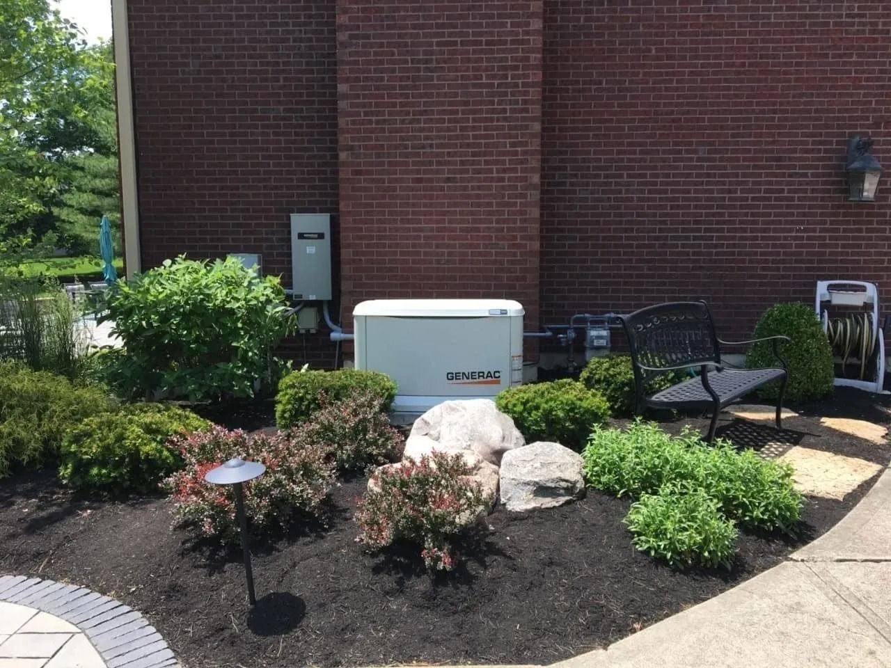 Brick building exterior with utility box, small landscaped shrubs, and patio chairs by a sidewalk
