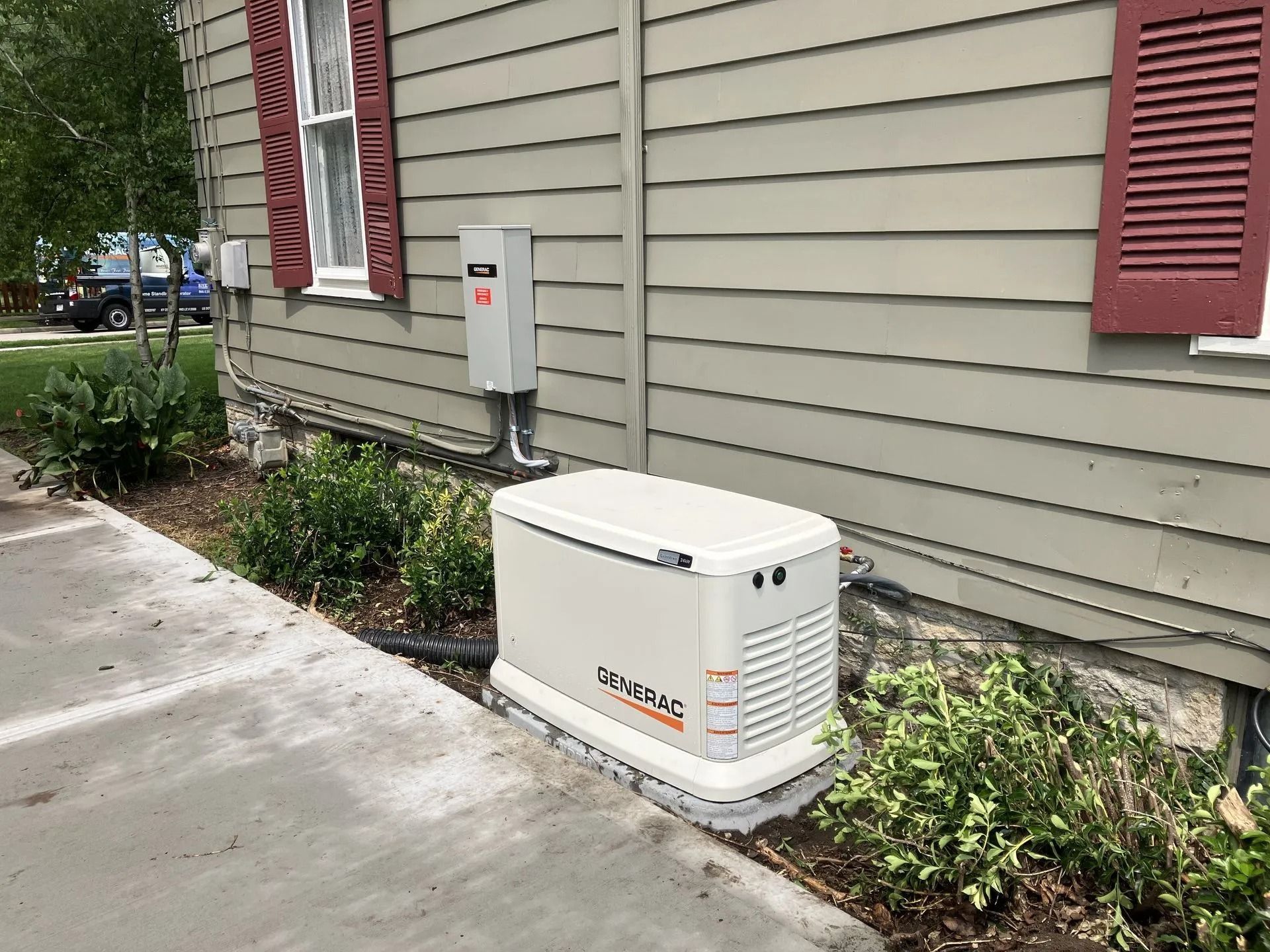 Outdoor generator beside a beige house, next to a driveway and shrubs