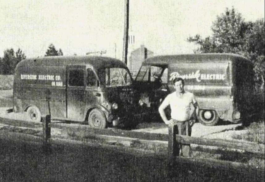 Two vintage electric delivery vans parked roadside, with a man standing in front