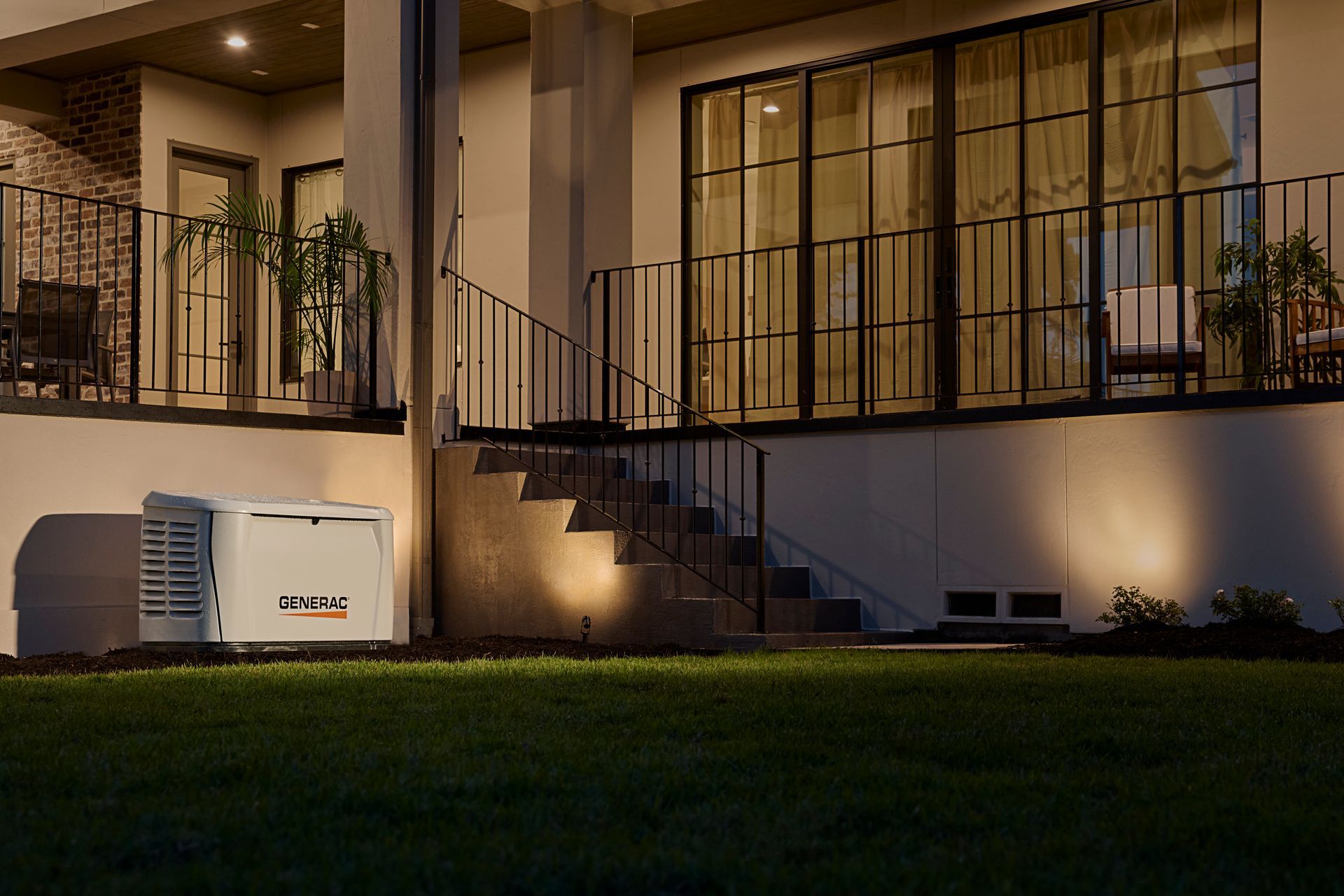 Nighttime porch and balcony of a house with warm lights, black railings, and a generator on the lawn.