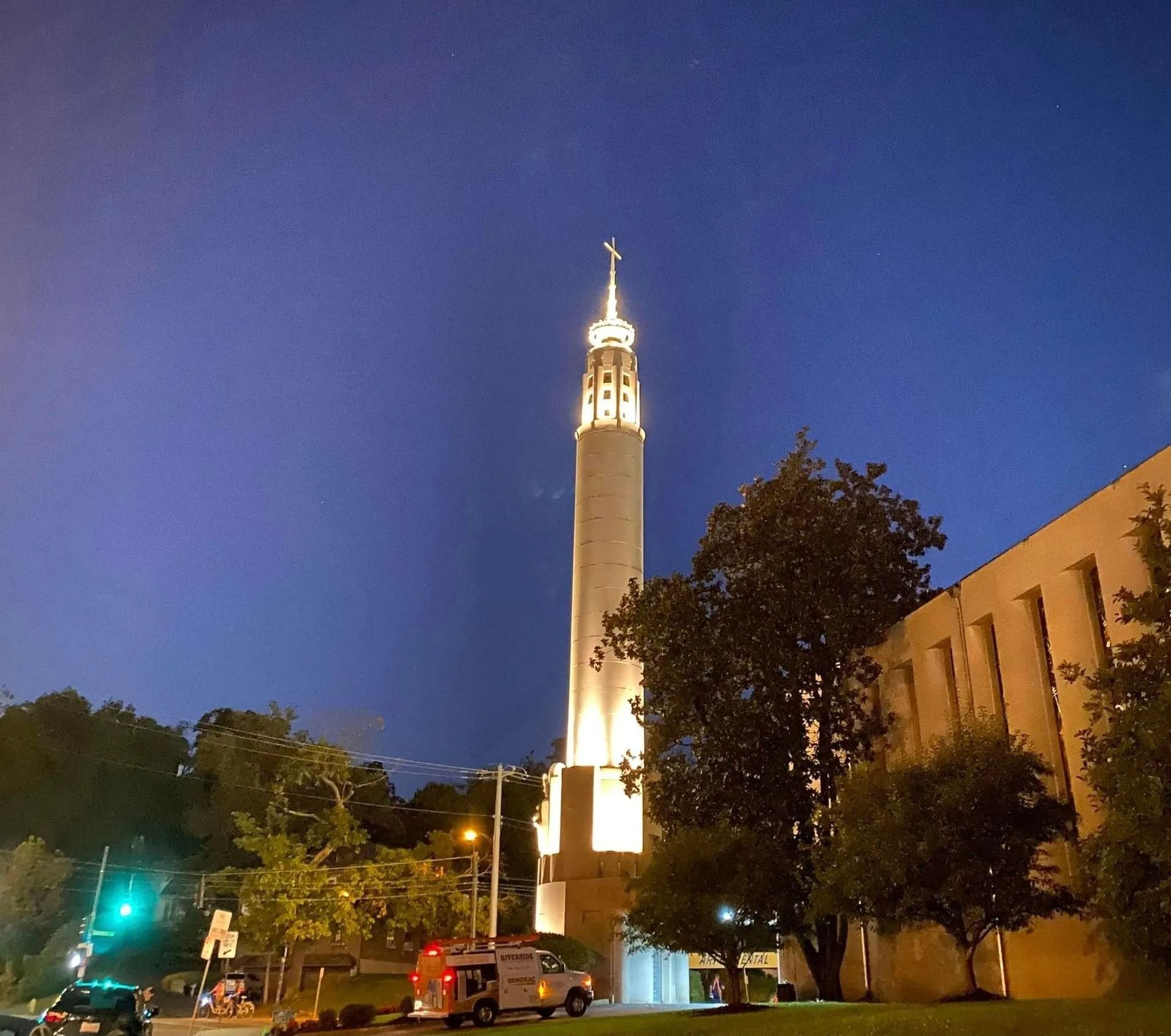 Illuminated church tower at dusk beside a building, with trees and parked cars in the foreground