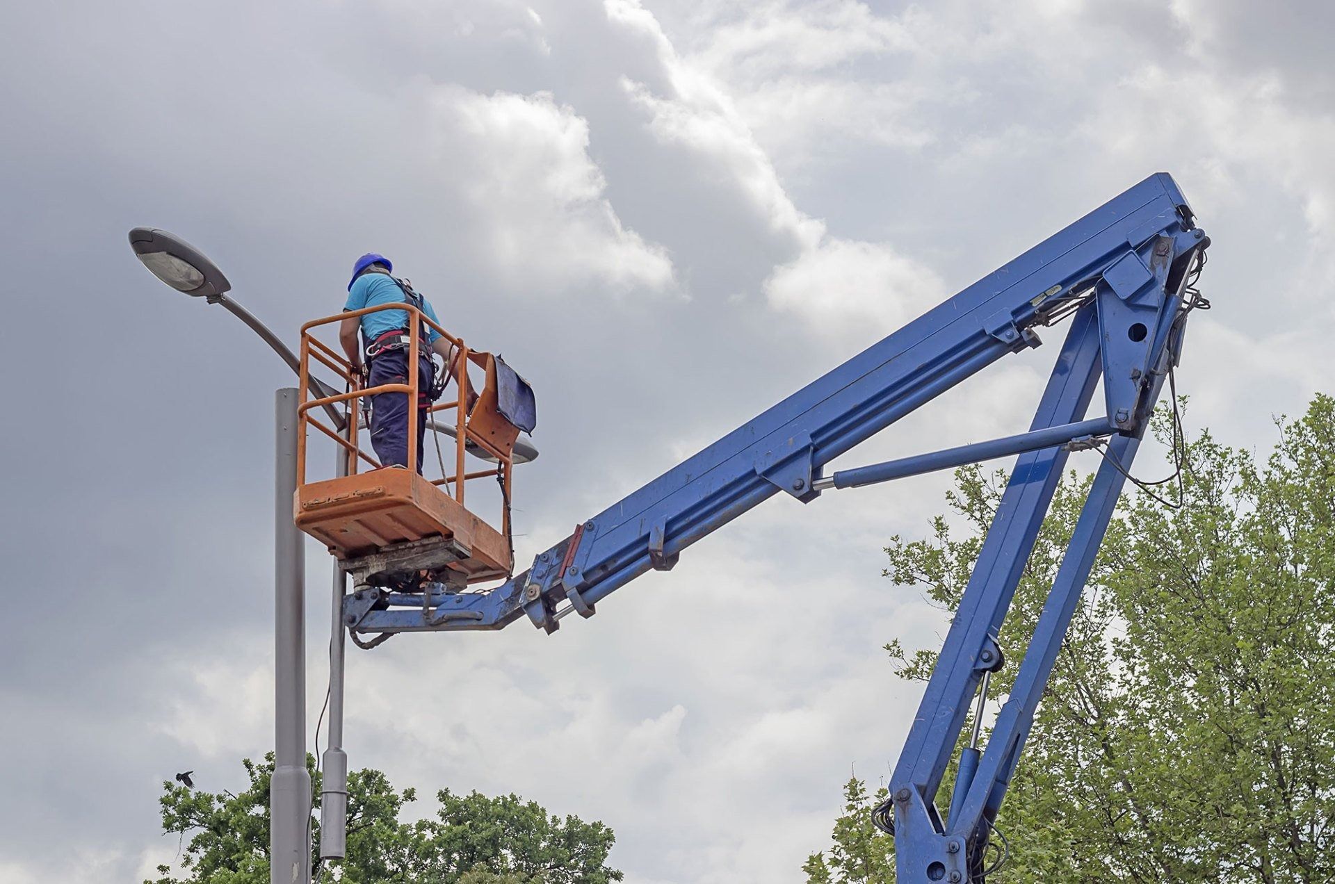 Worker in a blue lift basket raised beside a streetlight, with a blue crane boom against a cloudy sky