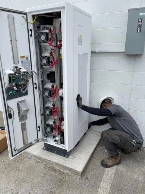 Technician crouches by an open electrical cabinet, inspecting equipment with a flashlight.