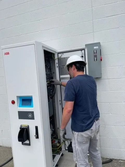 Technician in hard hat inspecting an open electrical control cabinet next to a wall-mounted panel.