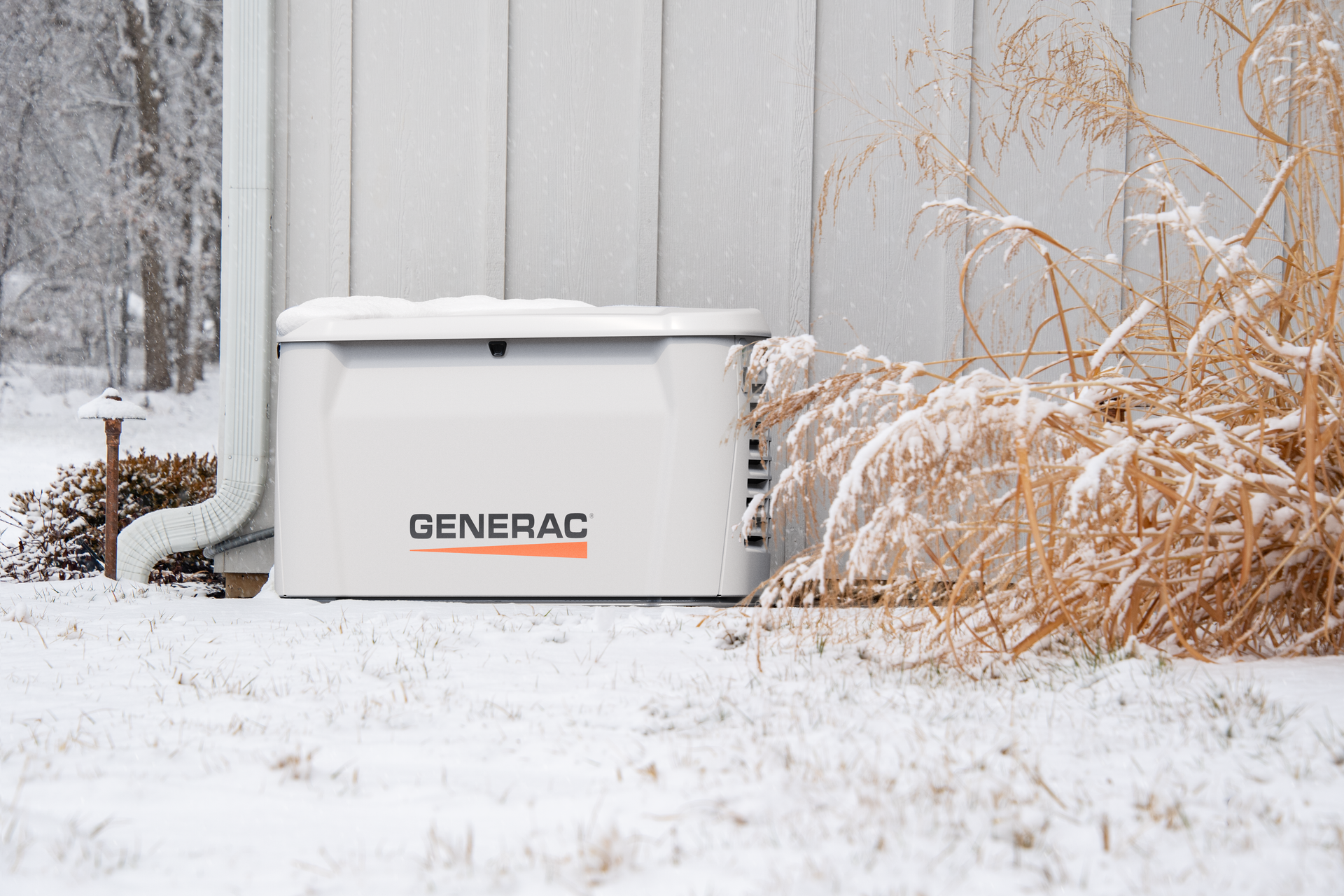 White Generac backup generator beside a house in a snowy yard with dried plants nearby