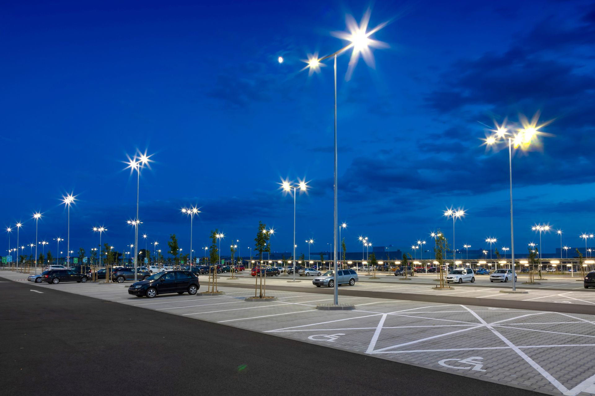 Nighttime parking lot with bright streetlights and a few parked cars under a blue sky