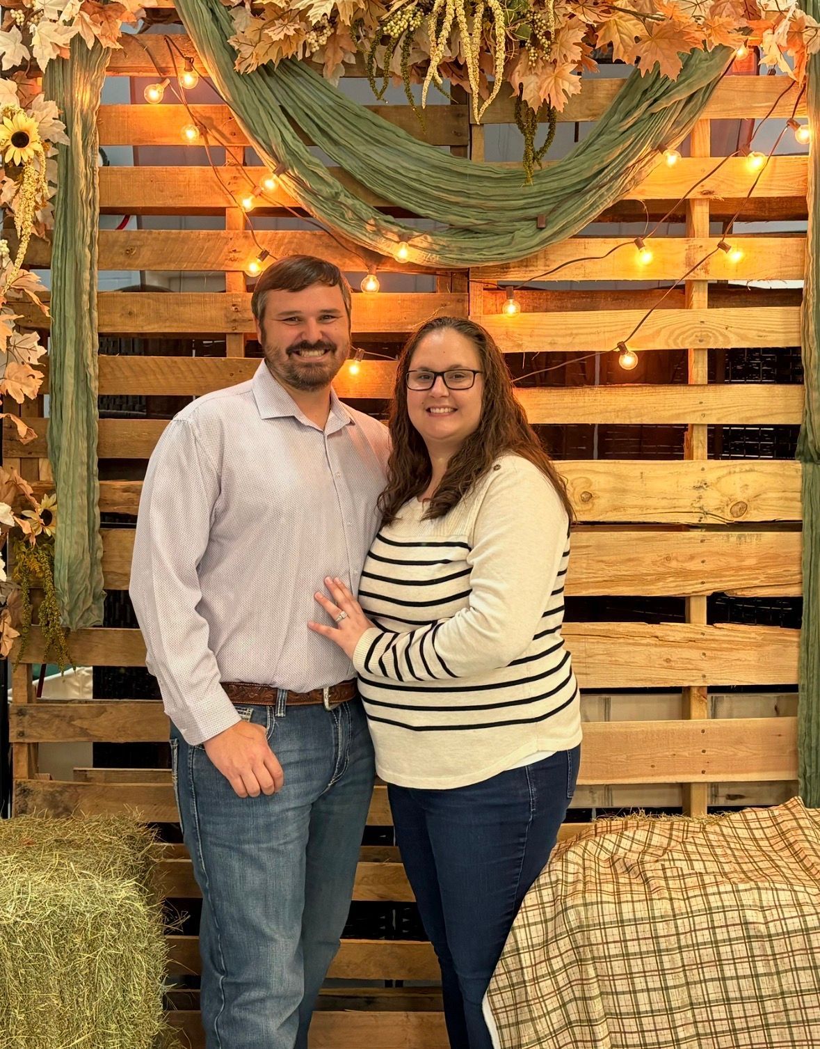 Couple smiling in front of a rustic wood backdrop with autumn leaves and string lights