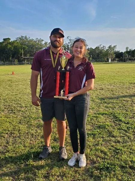Man and woman in maroon shirts hold trophy on a grassy field.