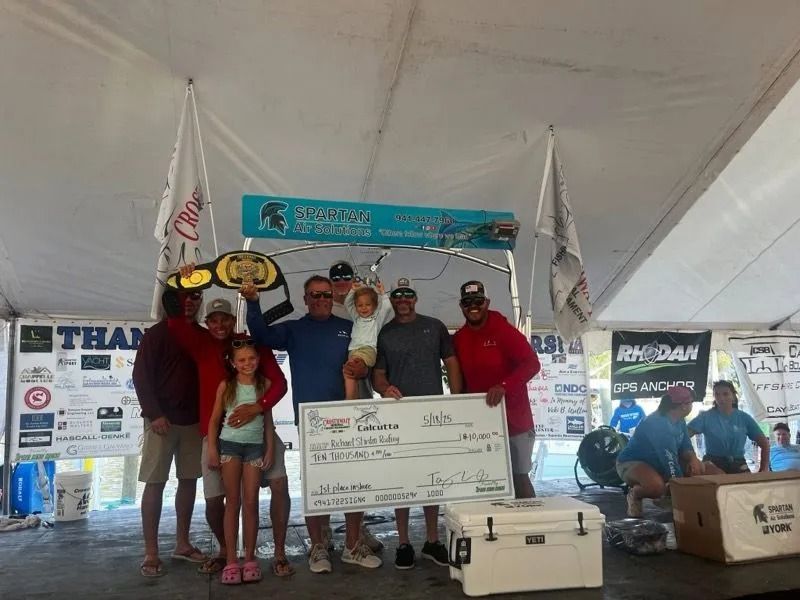 People celebrating a fishing tournament win under a tent; winner holding a large check and trophy belt.