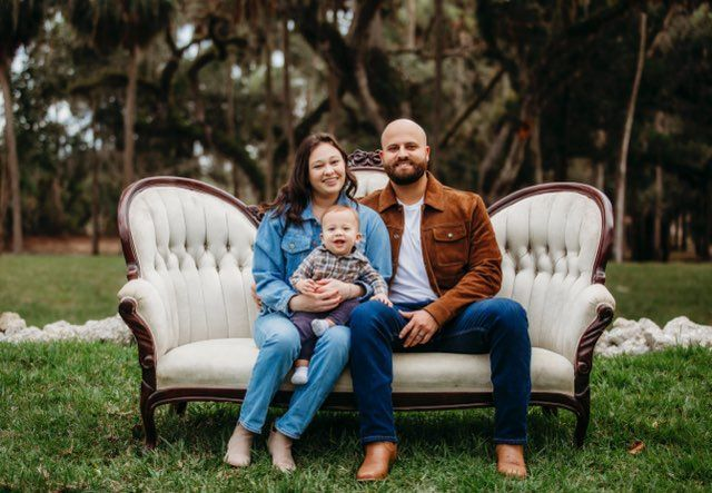 Family of three sits on an antique white couch outdoors; woman in denim, man in brown jacket, baby in arms.