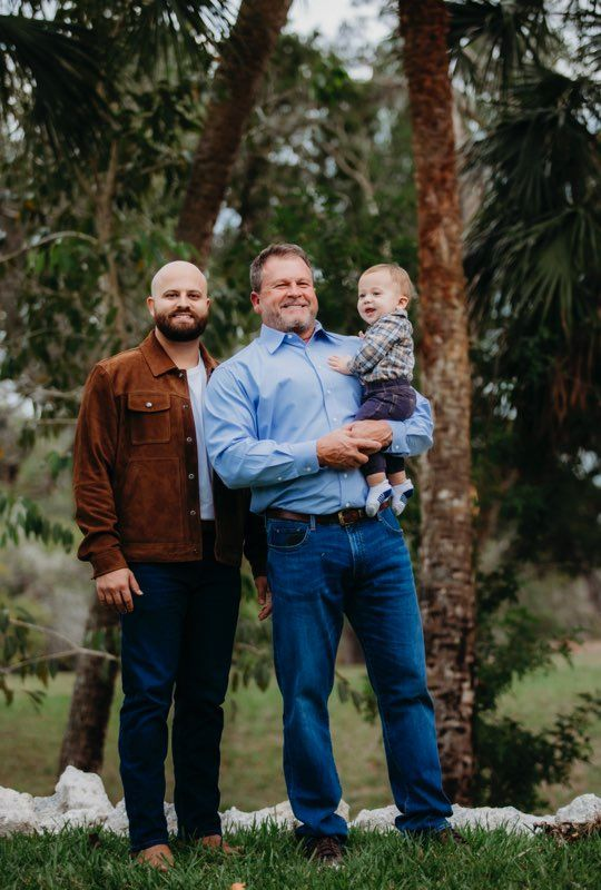 Two men and a baby in a forest setting. One man holds the baby; all smile.
