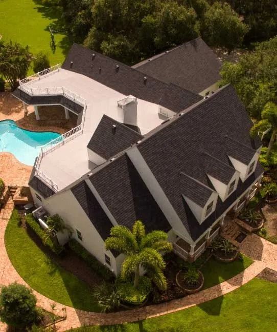 Aerial view of a white house with a black roof, a pool, and a green lawn.