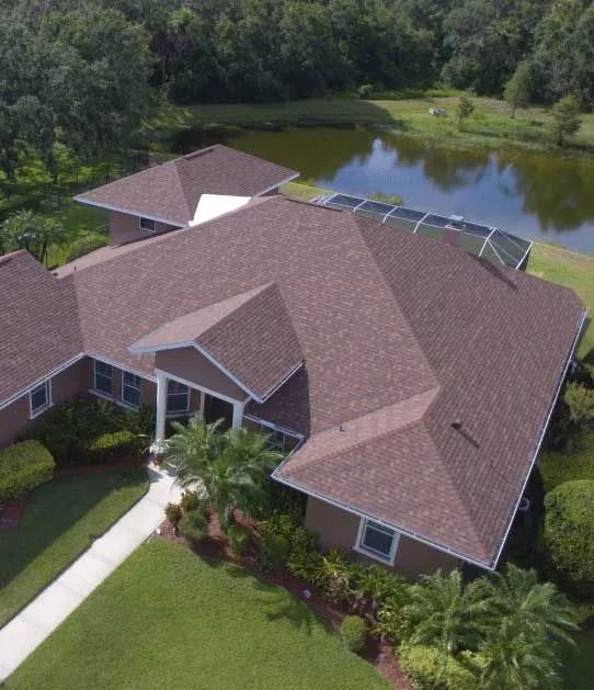 Brown-roofed house with a lake in the background, lush greenery, and a walkway leading to the entrance.