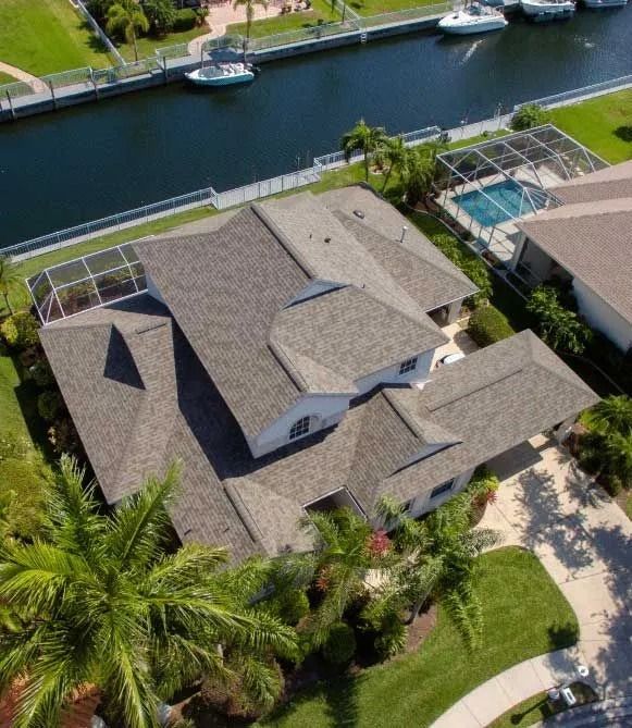 Aerial view of a house with a gray roof, next to a canal, with boats and palm trees.