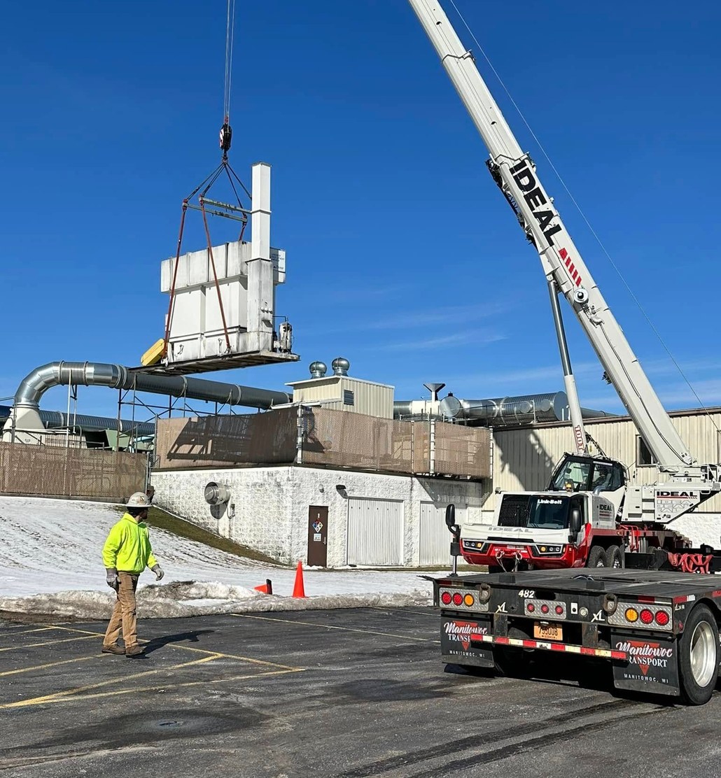 Crane lifting rooftop equipment on a snowy construction site under a blue sky