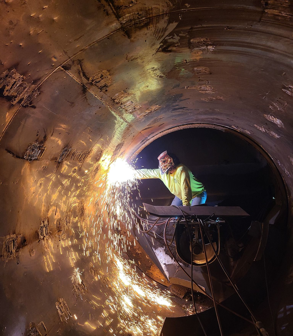 Worker in a neon vest grinding inside a large metal pipe, with sparks flying around.