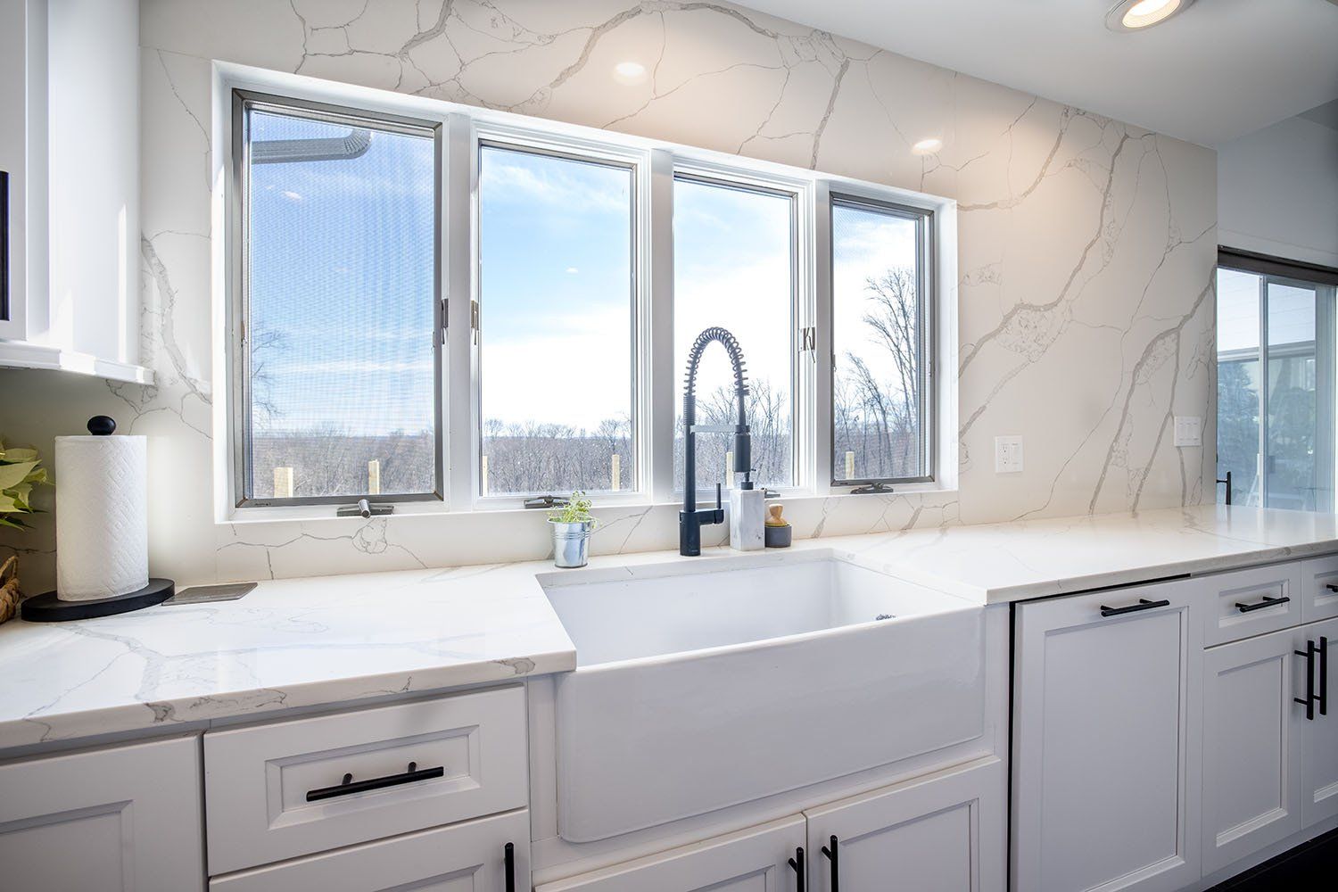 A kitchen with white cabinets , a white sink , and a large window.