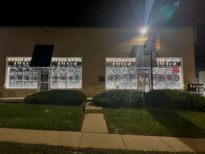Exterior of a store at night, with illuminated windows displaying products, a sign, and trimmed bushes in front.