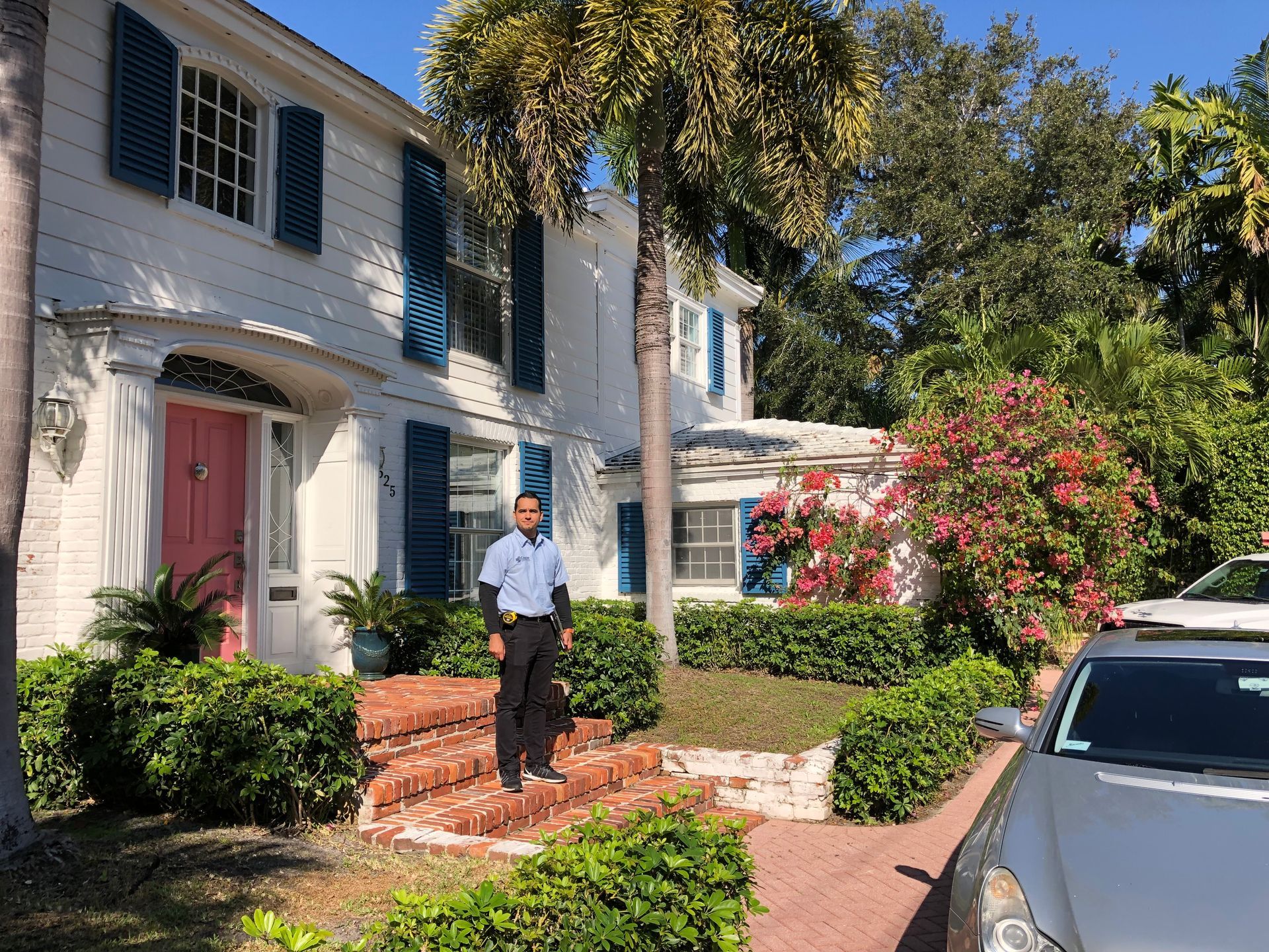 A man is standing in front of a large white house with blue shutters.