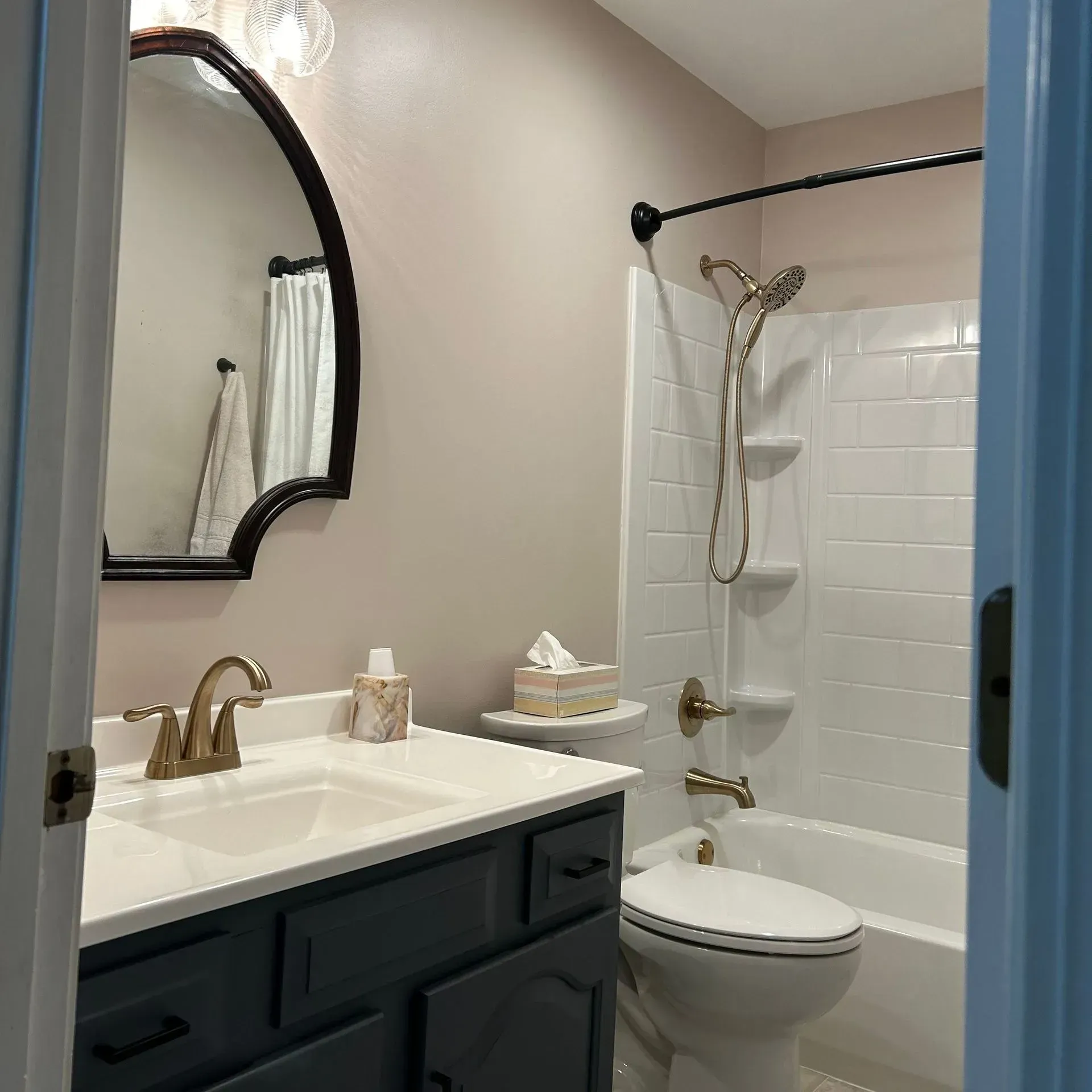 A bathroom with a dark vanity, white countertop, arched mirror, and a white tiled shower area with a handheld showerhead.