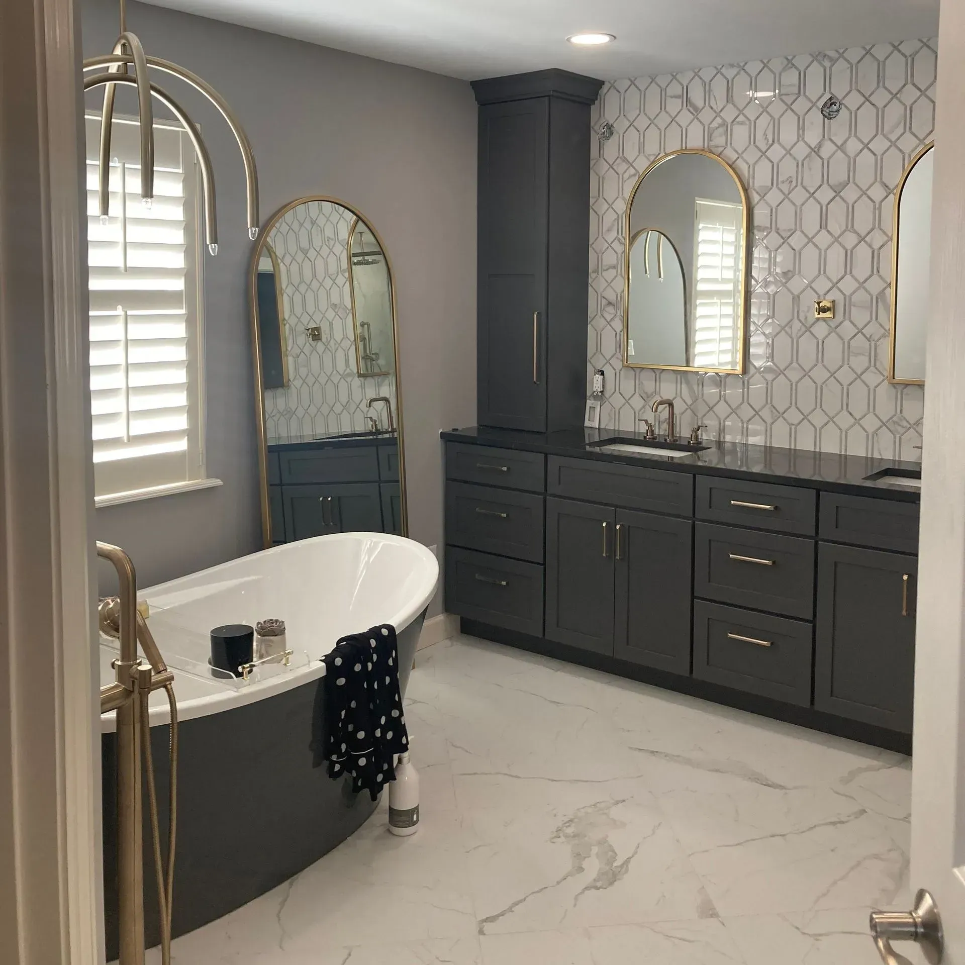 Modern bathroom featuring a charcoal vanity, soaking tub, arched gold-framed mirrors, and white marble-patterned flooring.