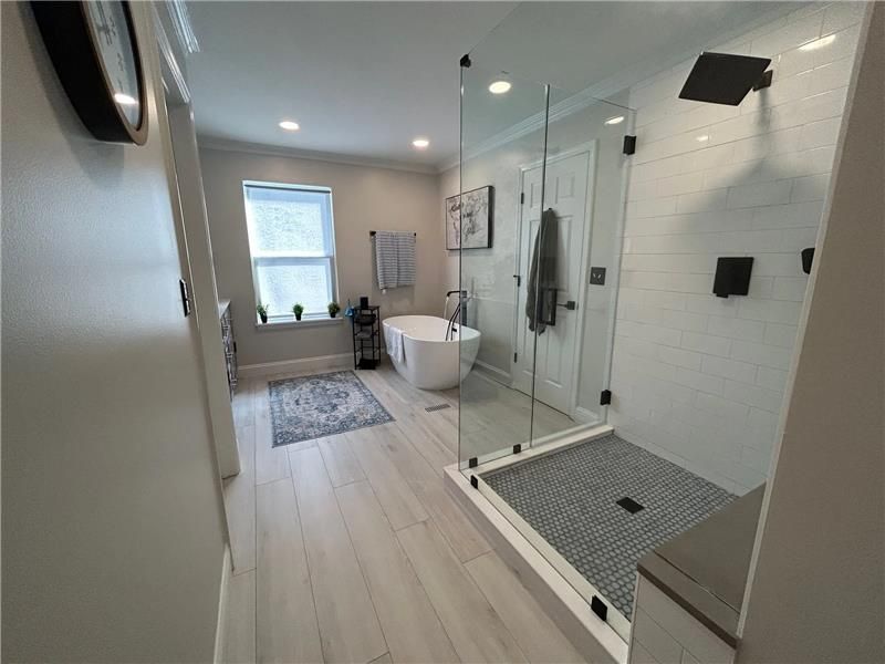 A modern, bright bathroom with light wood flooring, a glass-enclosed walk-in shower, and a freestanding white bathtub.