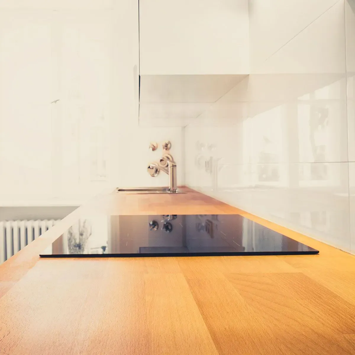 A modern kitchen counter with a light wood surface, a black induction cooktop, and a chrome faucet against white tiles.