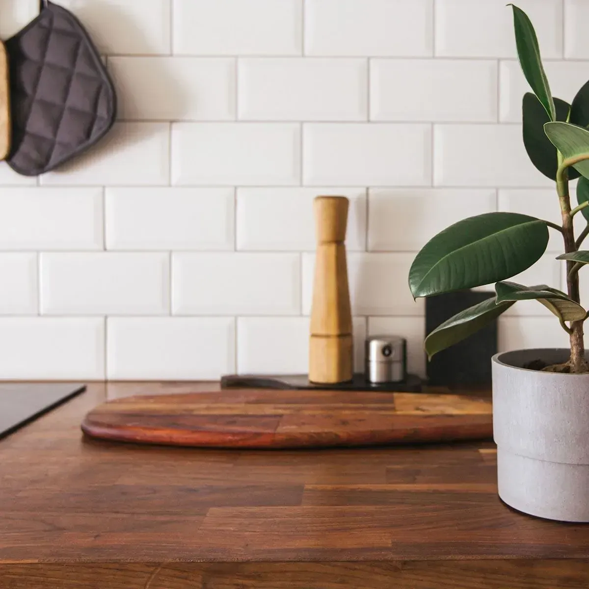 A wooden cutting board on a dark countertop with a pepper grinder and a potted rubber plant against a white tiled wall.