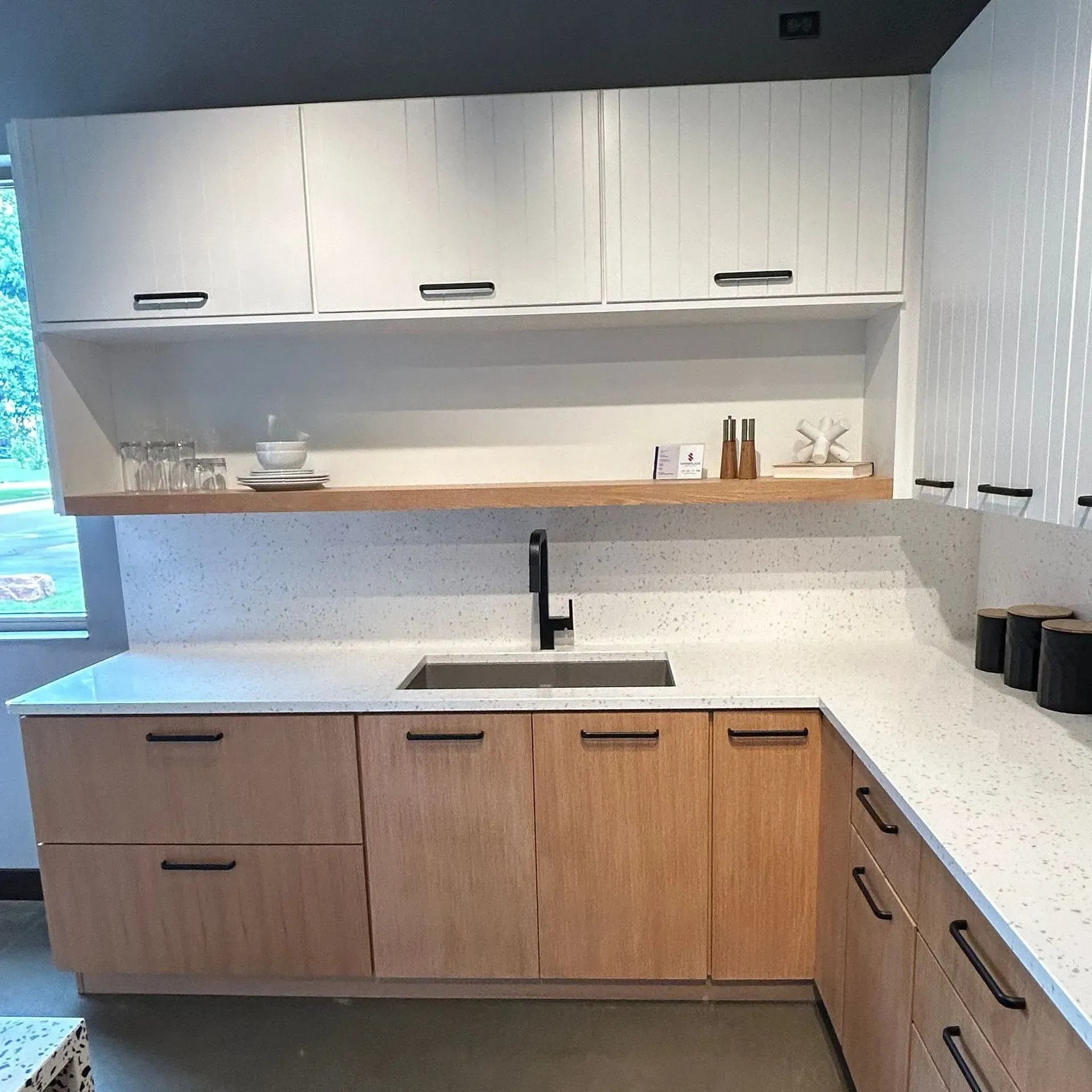 A modern kitchen with light wood lower cabinets, white upper cabinets, a white speckled countertop, and open shelving.